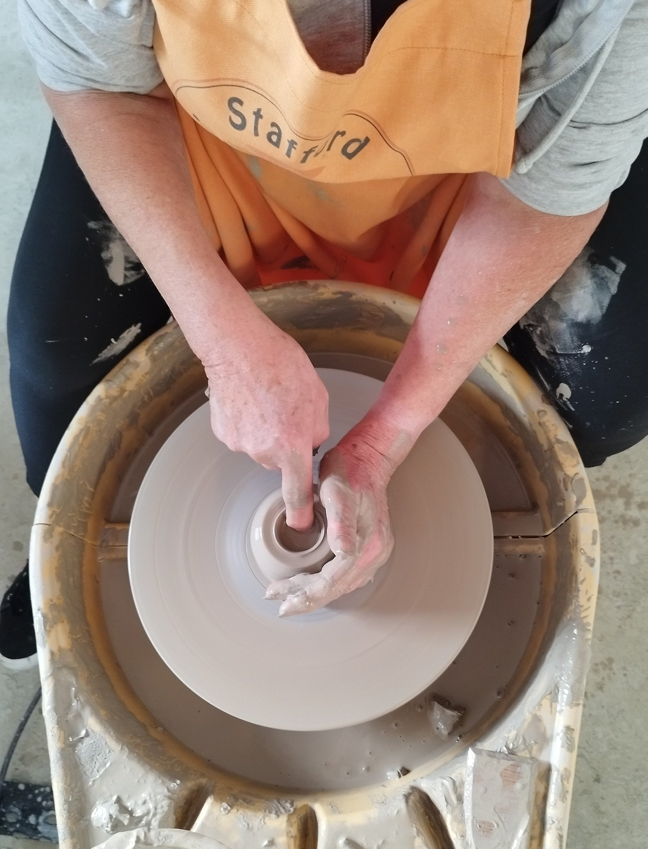 Person shaping clay on a pottery wheel, wearing an apron and using their hands to mold the clay.