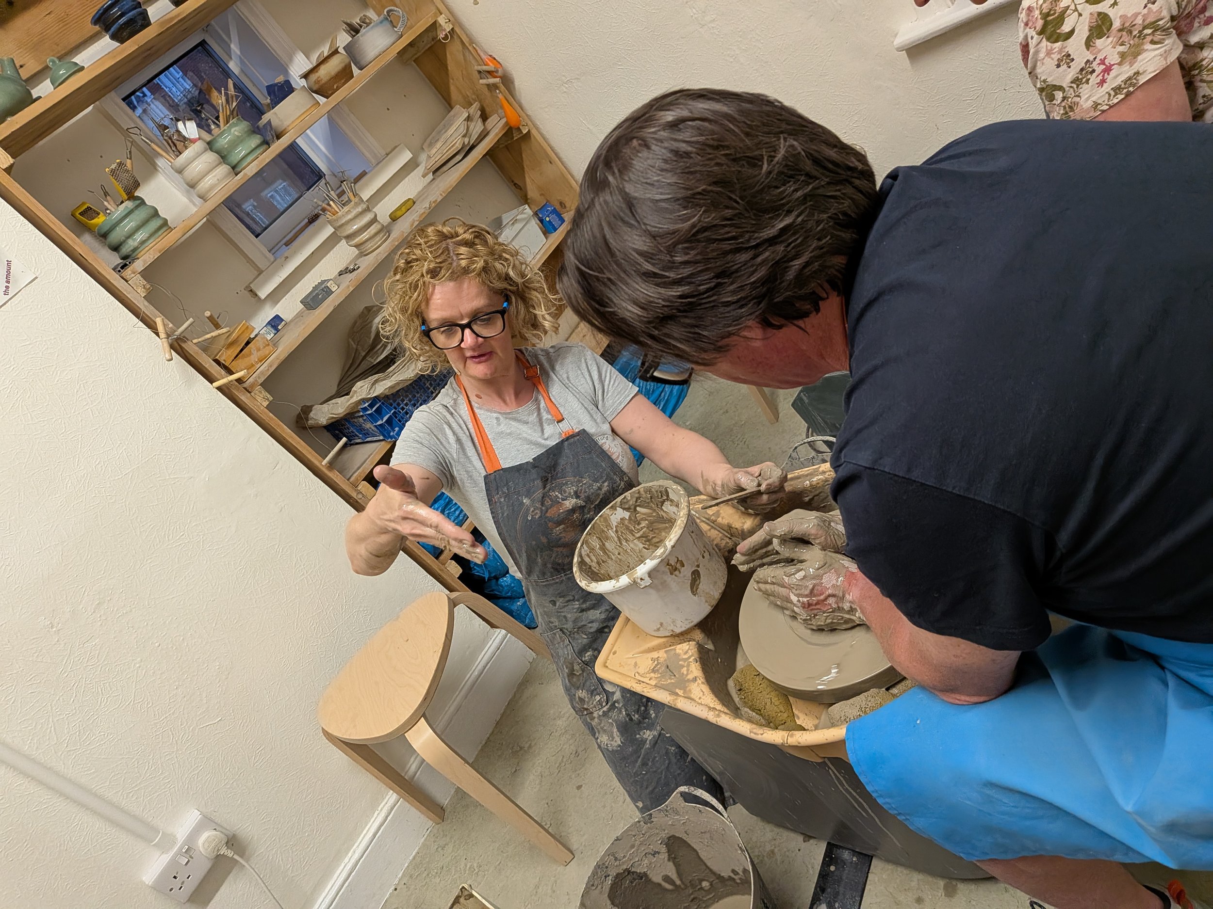 Two people in a pottery studio working on a ceramic piece using a pottery wheel. A woman with curly hair and glasses is instructing a man. Shelves with pottery items and tools are visible in the background.