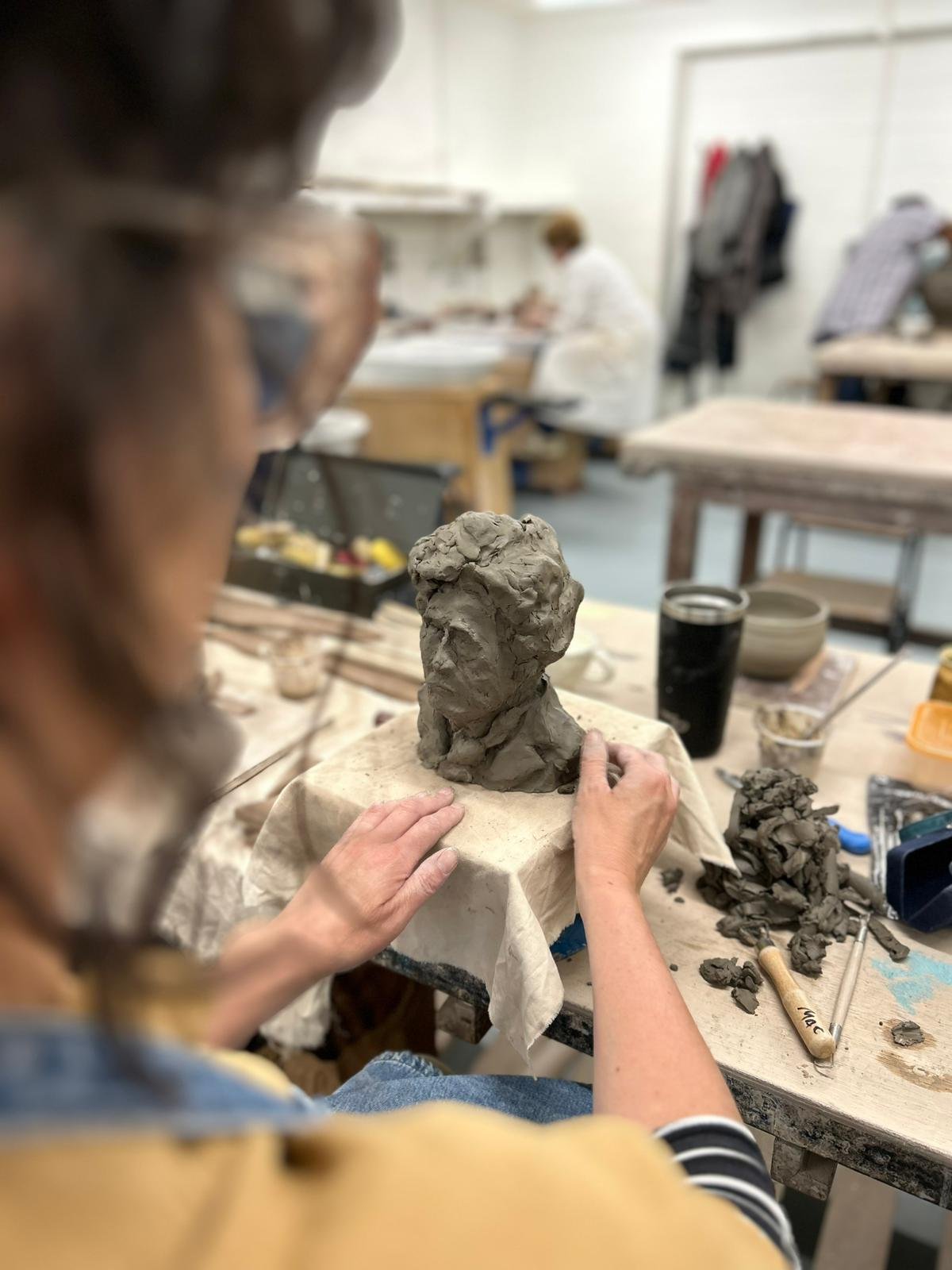 Person sculpting a clay bust on a table in an art studio, with clay tools nearby.