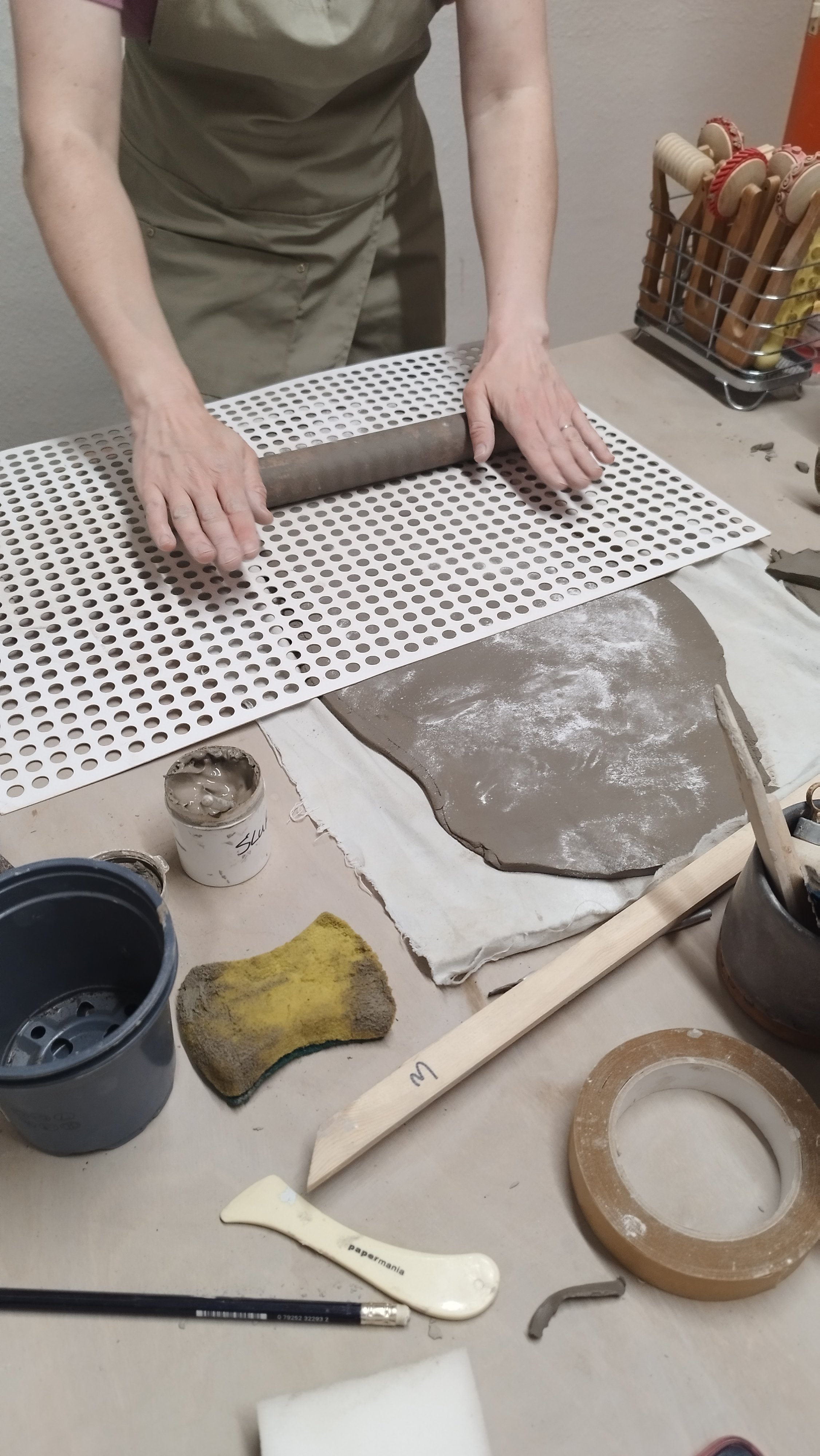 Person using a rolling pin on clay with tools and materials on a table in a pottery workshop.