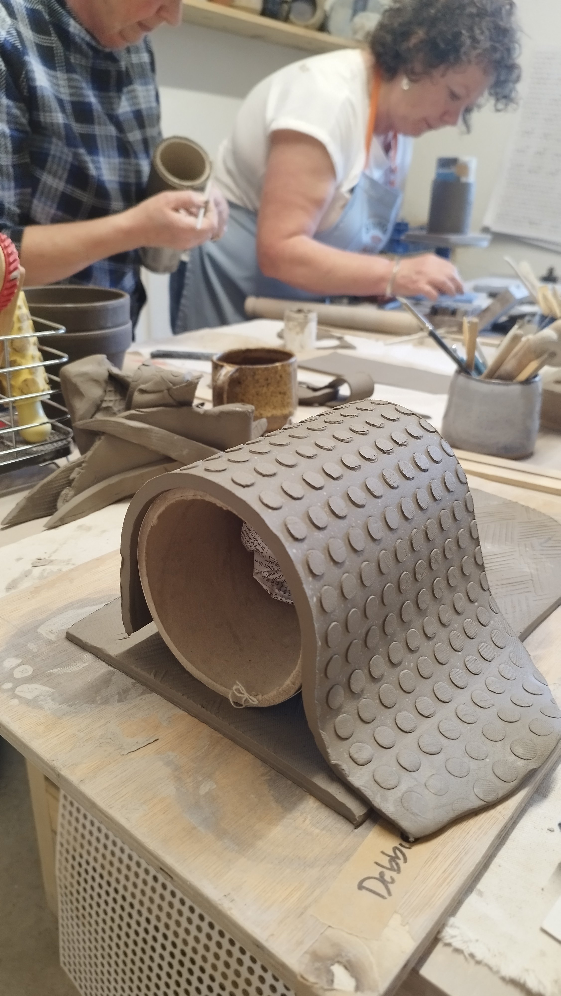 Two people working on pottery projects in a ceramics studio, with clay pieces and tools on a table.