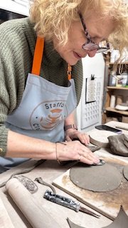 Person working with clay in a pottery studio, wearing glasses and an apron.