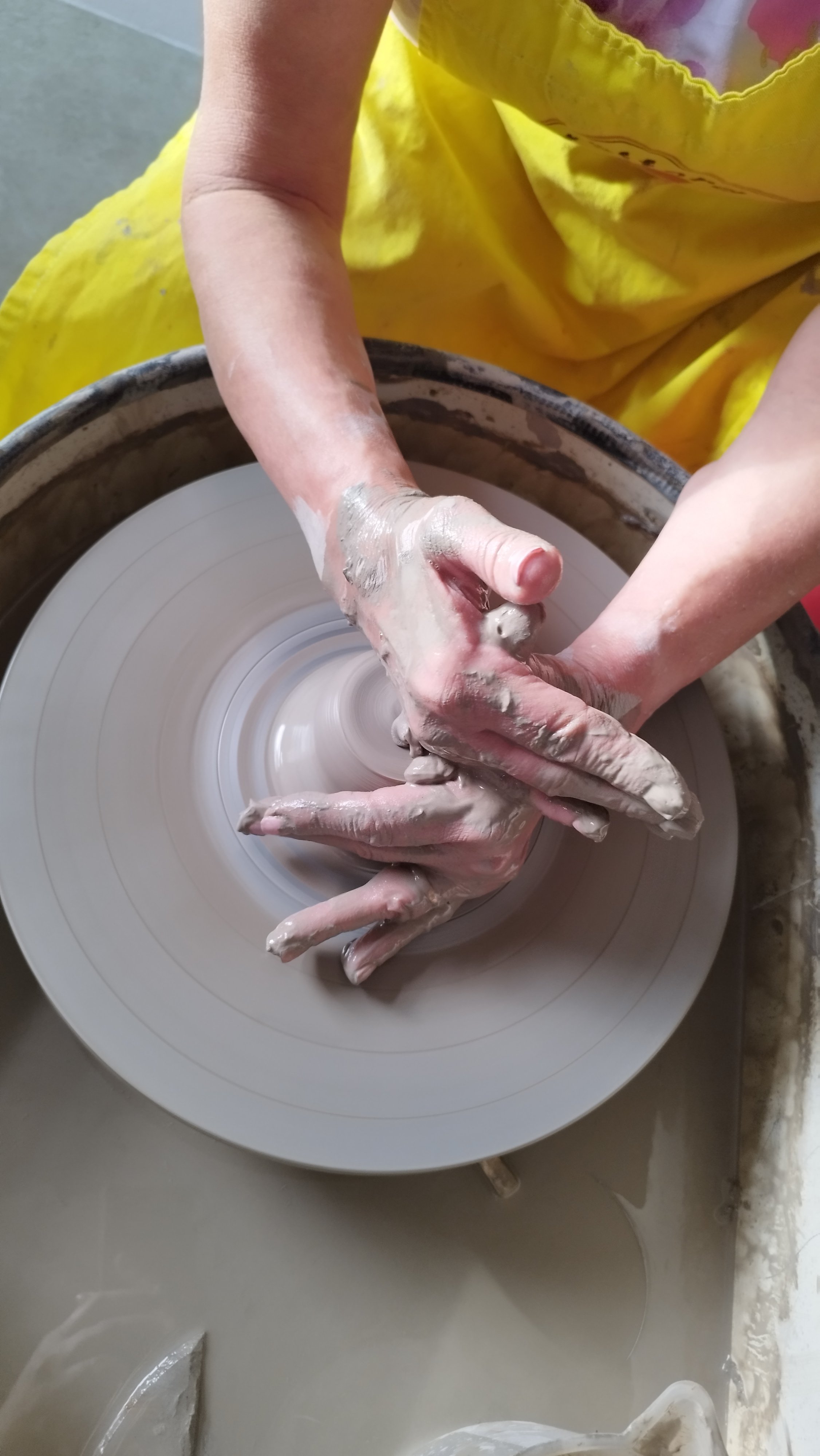 Hands shaping clay on a pottery wheel with a yellow apron in the background.
