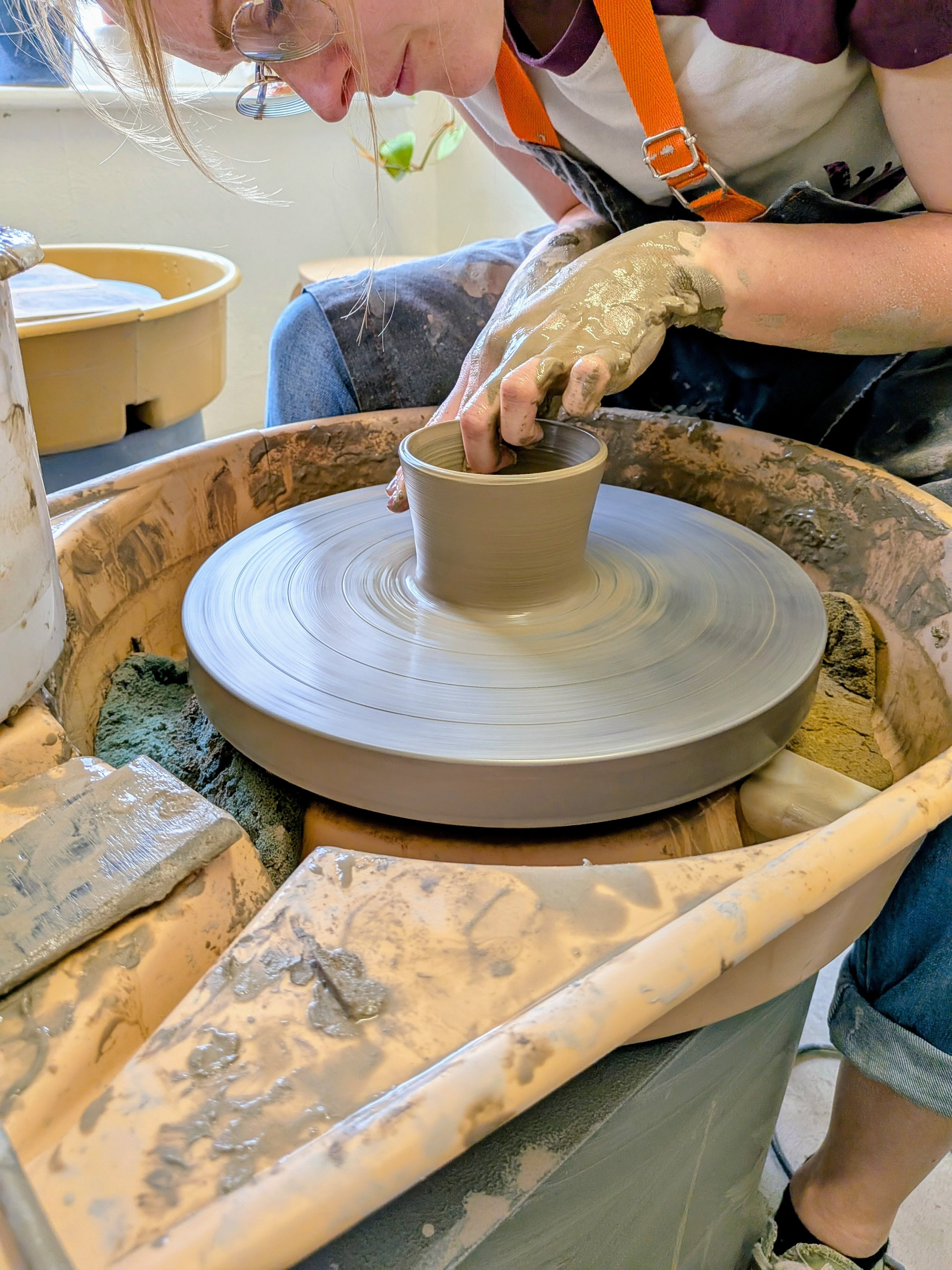 Person crafting clay pottery on a spinning pottery wheel, using hands to shape a small pot, with visible clay splatters.