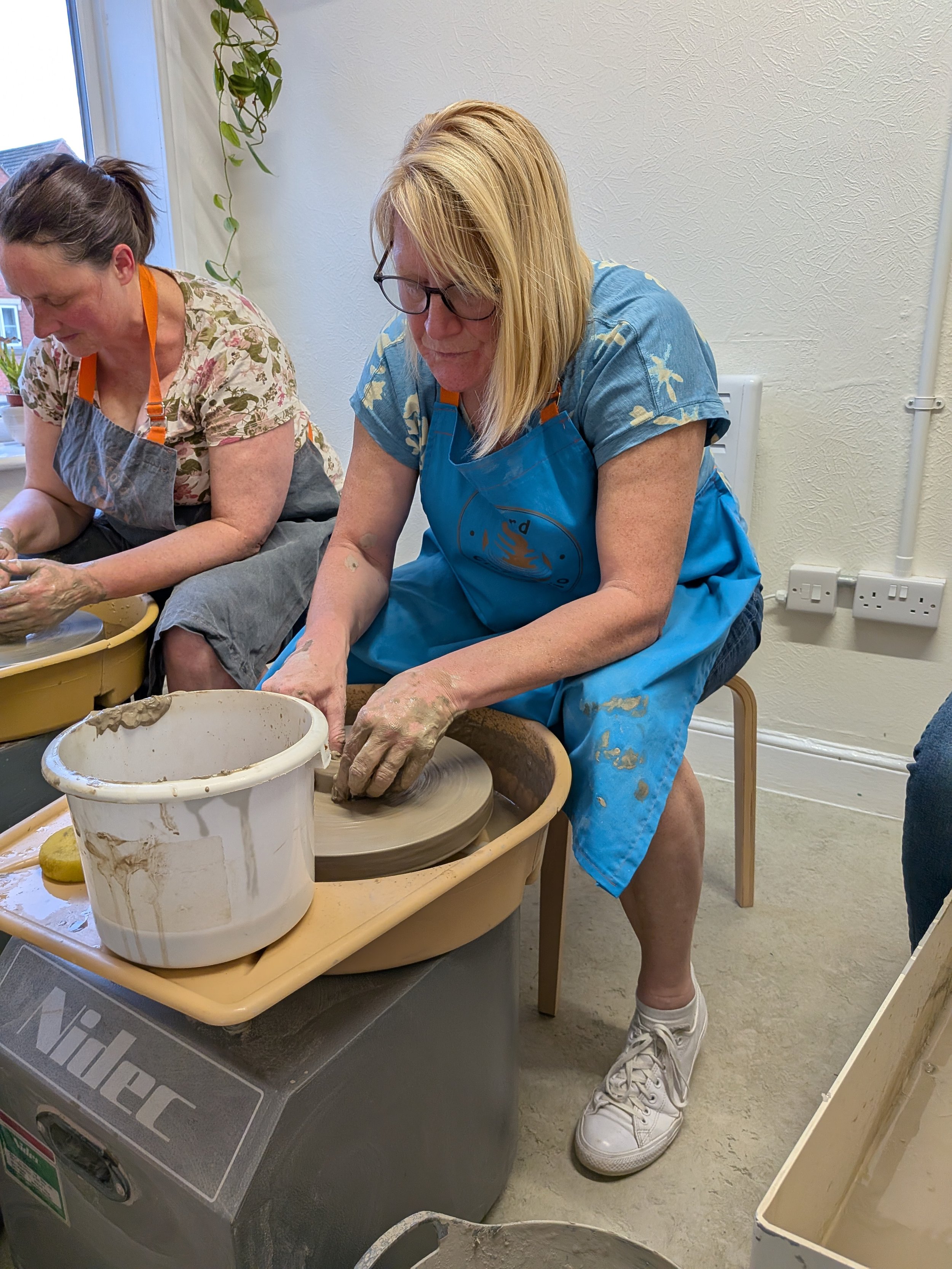Two women participating in a pottery class using pottery wheels and clay, wearing aprons, with tools and buckets nearby.