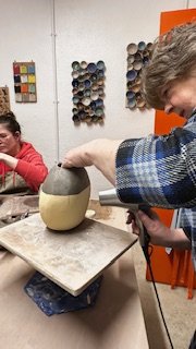 Two people engaged in pottery work, one using a heat tool on a ceramic piece, in a workshop with colorful clay samples on the wall.