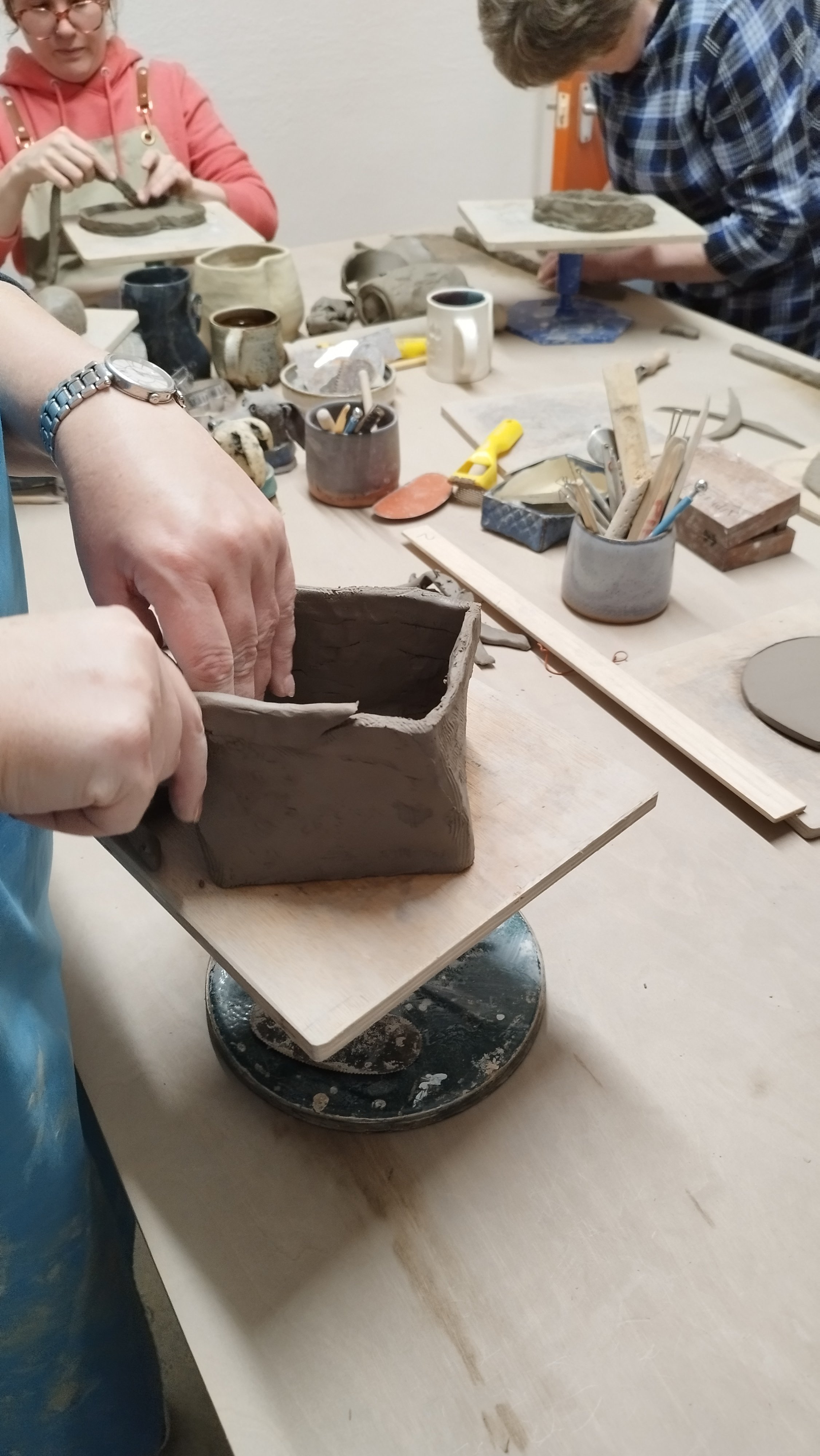 People working on pottery projects at a workshop table, with tools and materials scattered around. A person shapes clay into a square vessel on a rotating base.