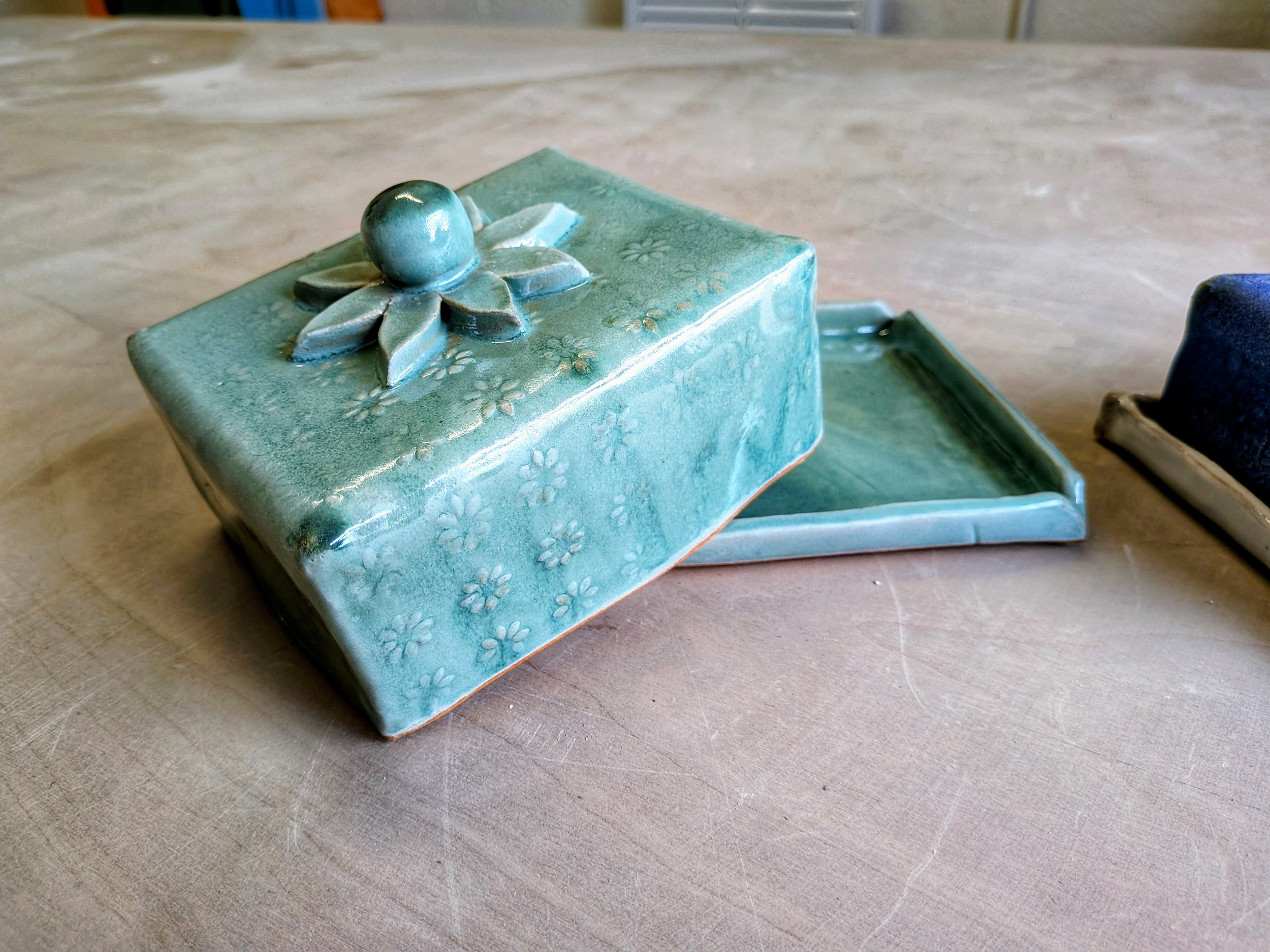 Handmade ceramic box with a lid featuring floral designs and a knob, on a table.