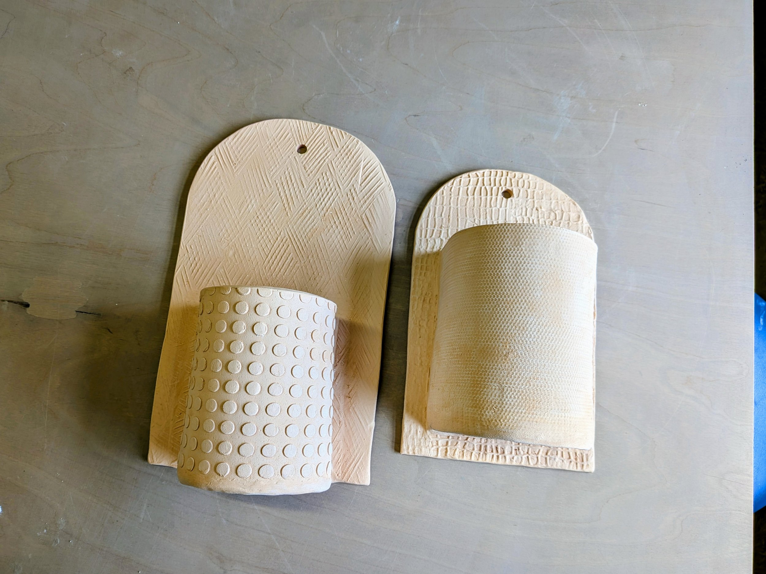 Two clay wall sconces with textured patterns, one with dots and the other with a fabric-like texture, placed on a wooden surface.