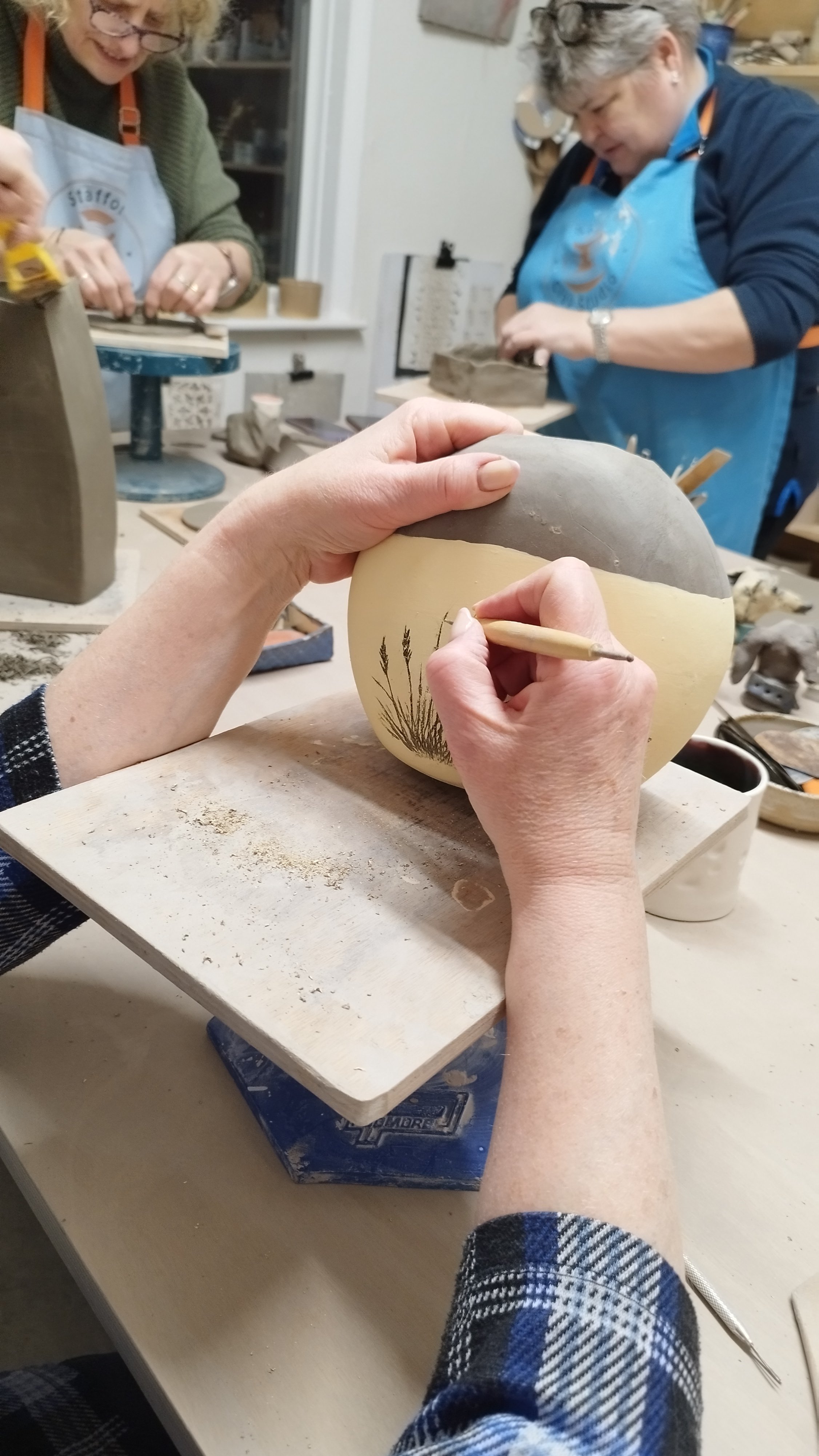 People working on pottery projects in a crafts workshop, with one person engraving a plant design on a round clay piece.