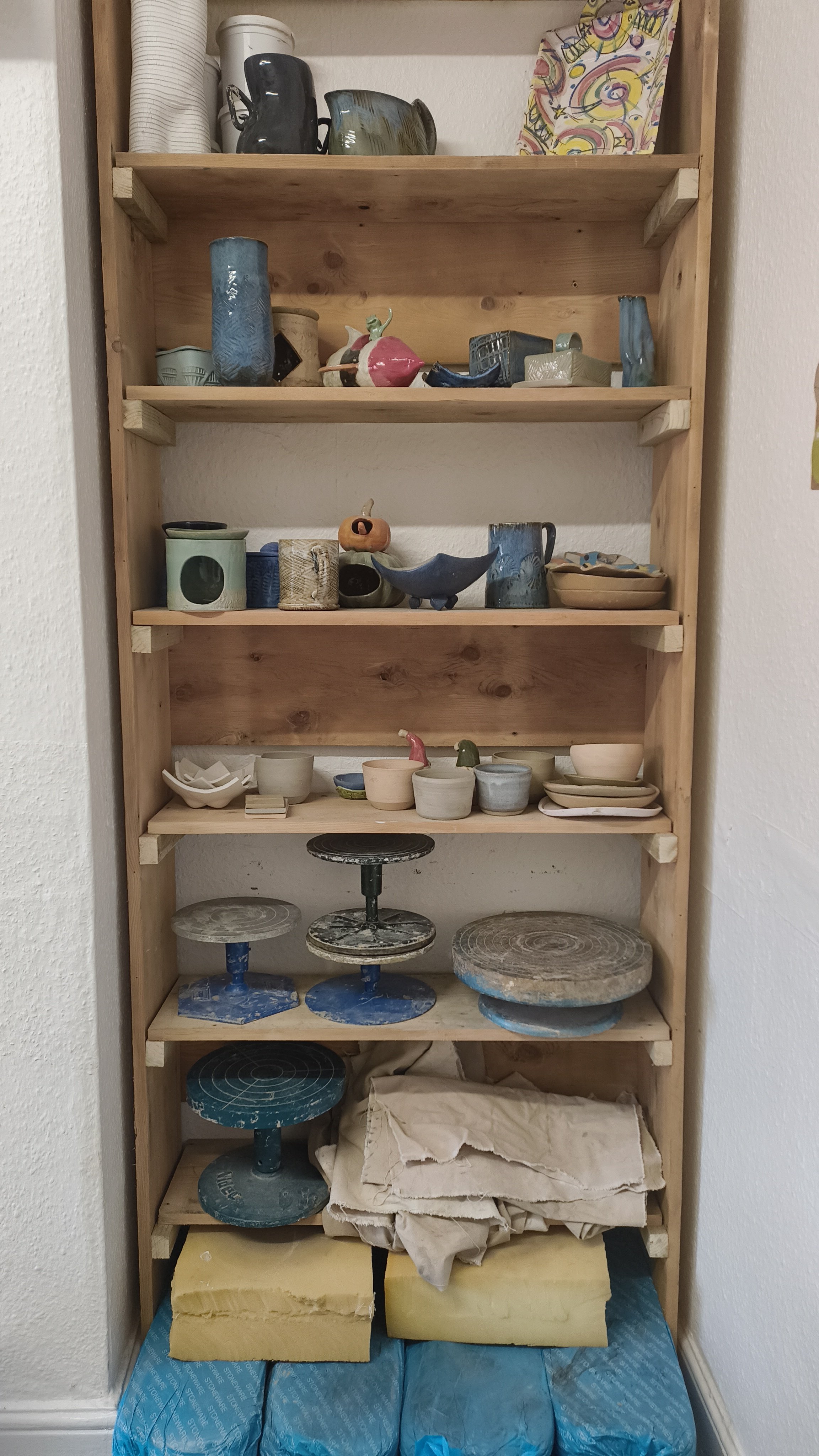 Wooden shelf with assorted pottery and ceramic items, art supplies, and bags of clay