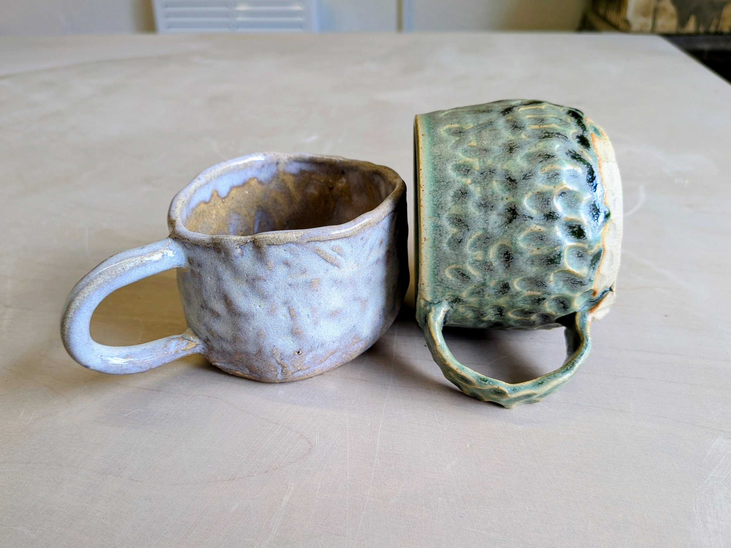 Two handmade ceramic cups with textured surfaces; one is light gray and the other is green.
