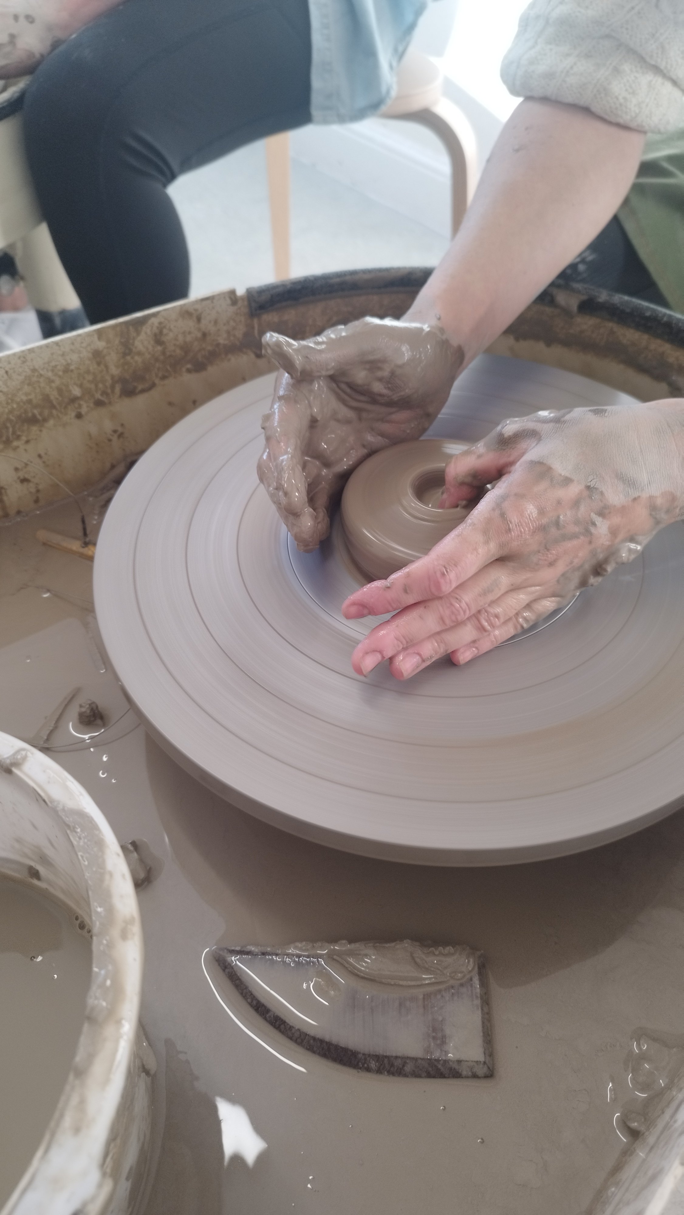 Clay pottery being shaped on a spinning wheel by hands; a pottery tool and bowl with clay slip are nearby.