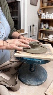 Person shaping clay on a pottery wheel in a studio surrounded by shelves with pottery supplies.