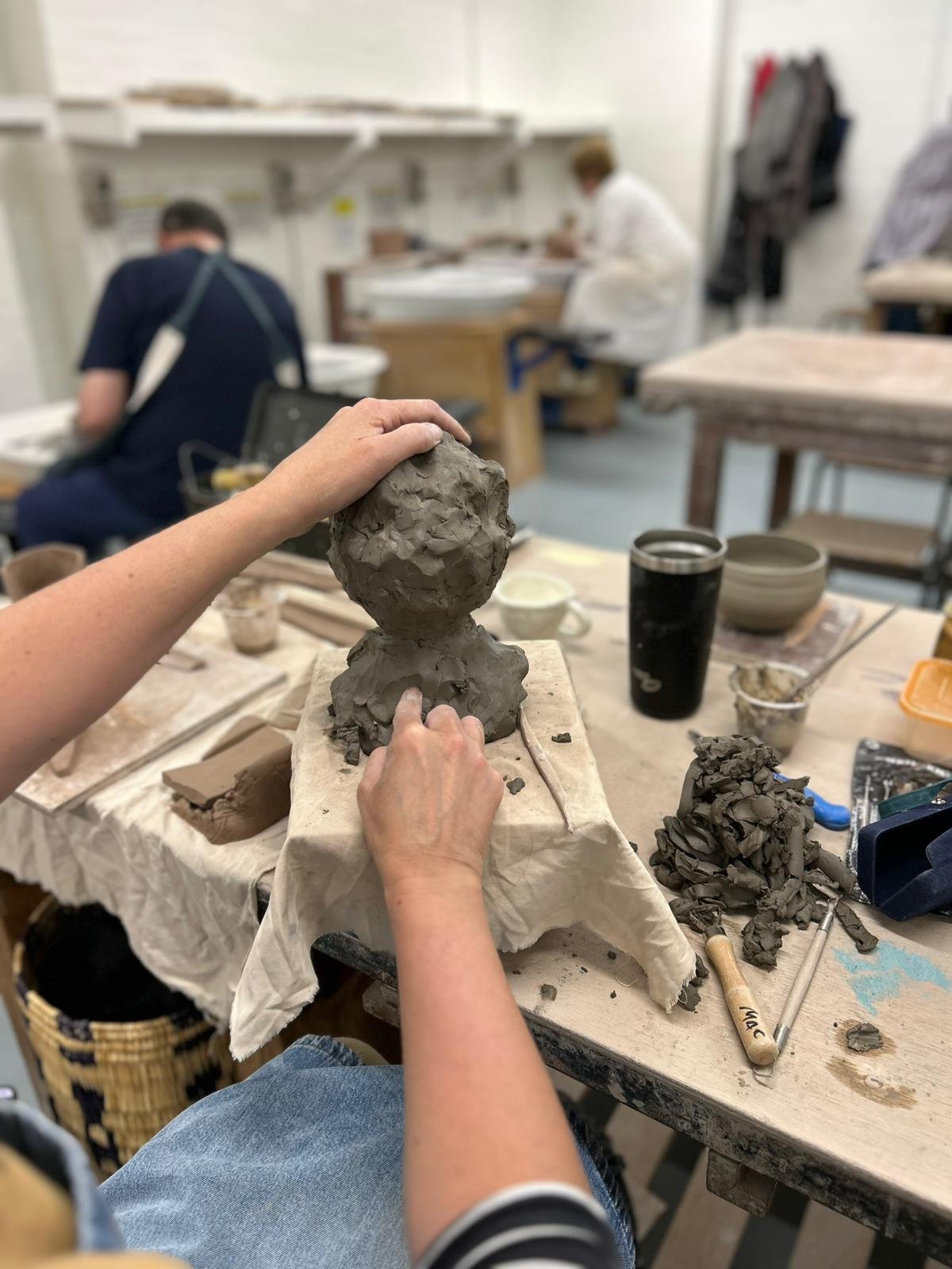 Person sculpting clay figure in a ceramics workshop, with tools and materials on the table.
