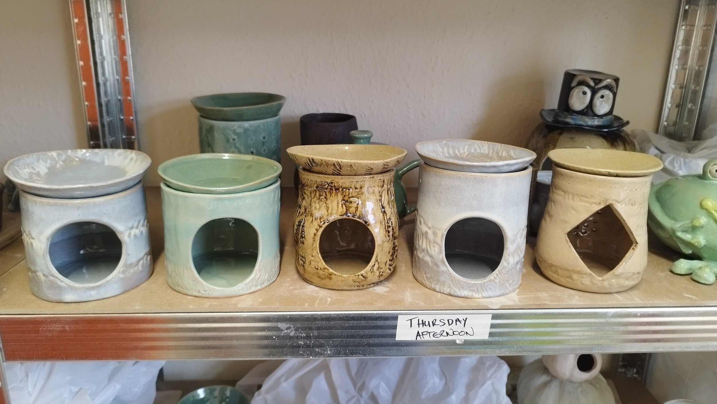 A shelf displaying five ceramic oil burners in various colors and designs, alongside other pottery and labeled 'Thursday Afternoon.'