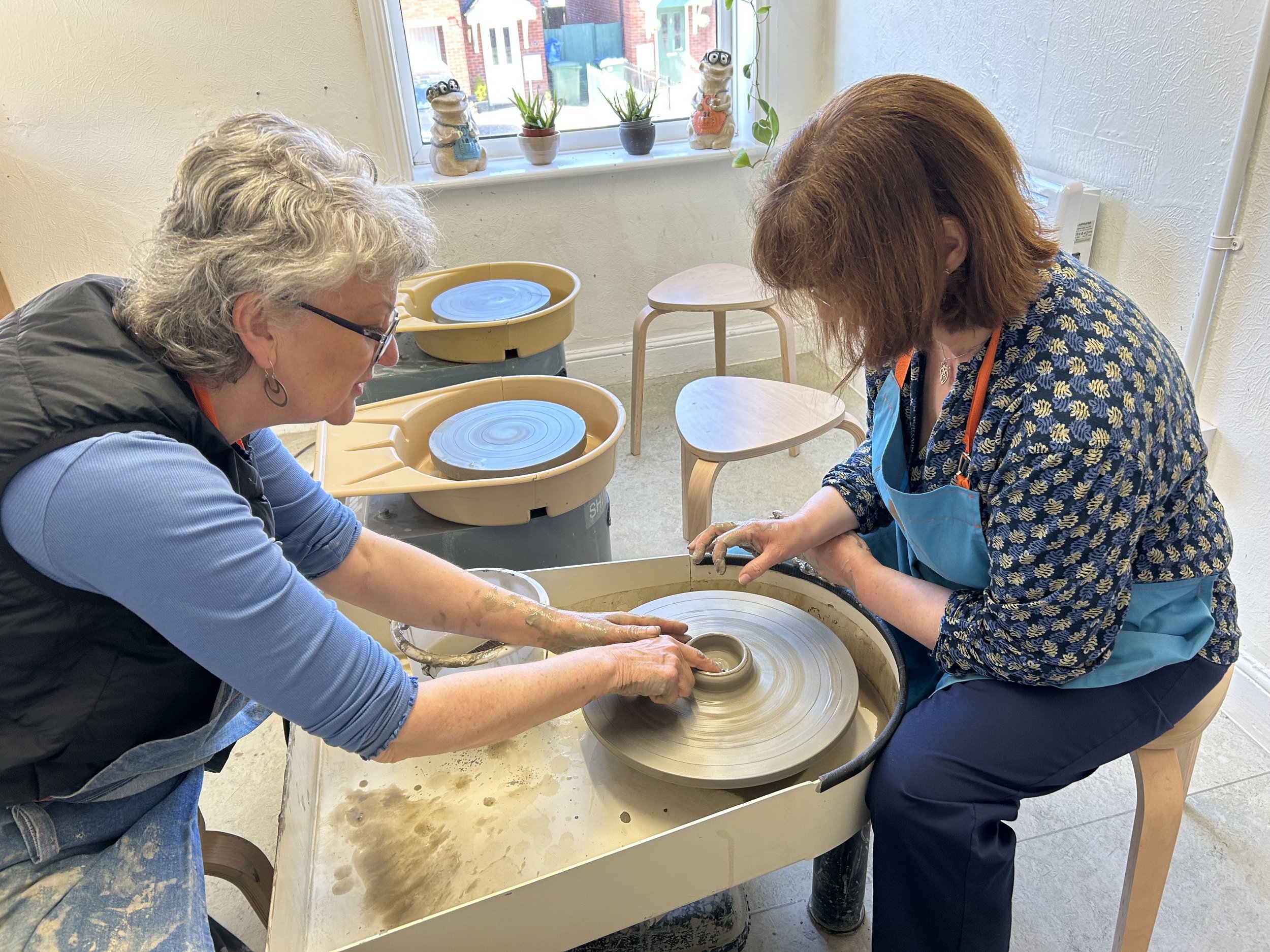 Two women working at a pottery wheel in a studio, shaping clay. One is instructing the other. The studio has a bright window with potted plants on the sill.