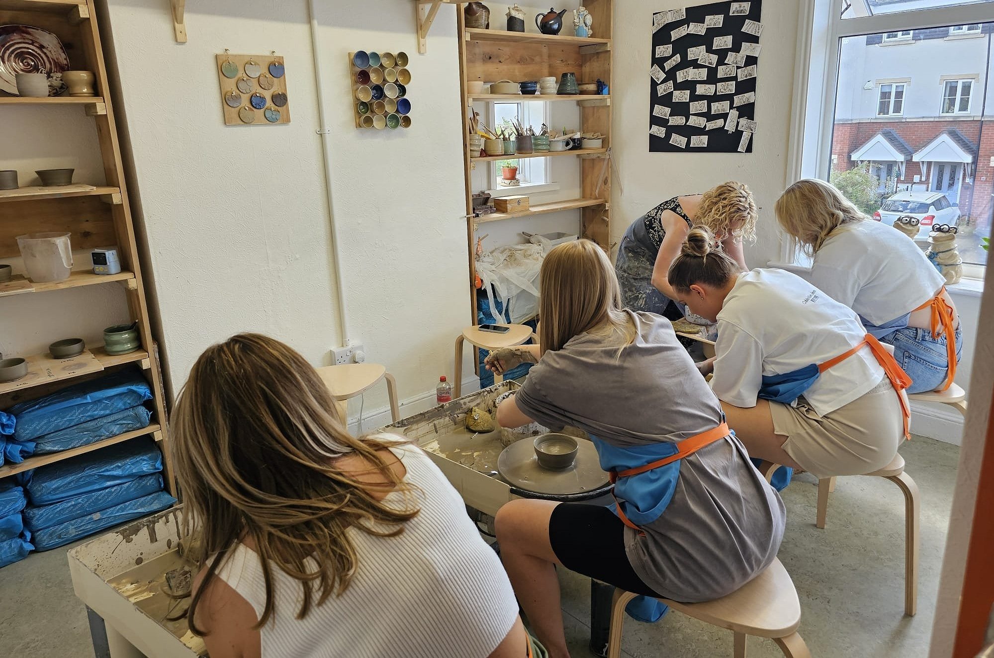 People participating in a pottery class, using pottery wheels to shape clay, in a room with shelves of ceramics and art materials.