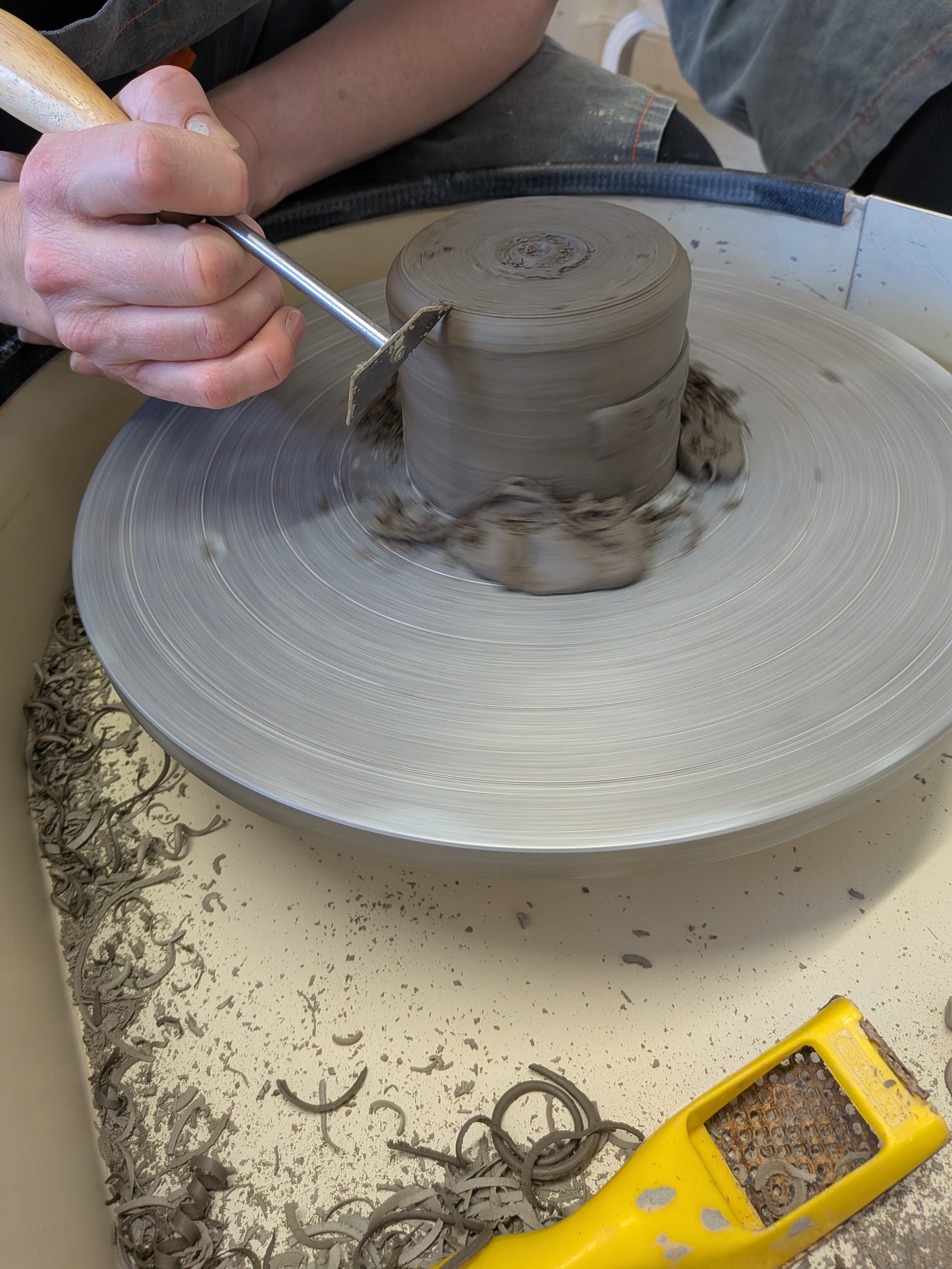 Person shaping clay on a pottery wheel with a trimming tool, surrounded by clay shavings and a yellow pottery tool nearby.