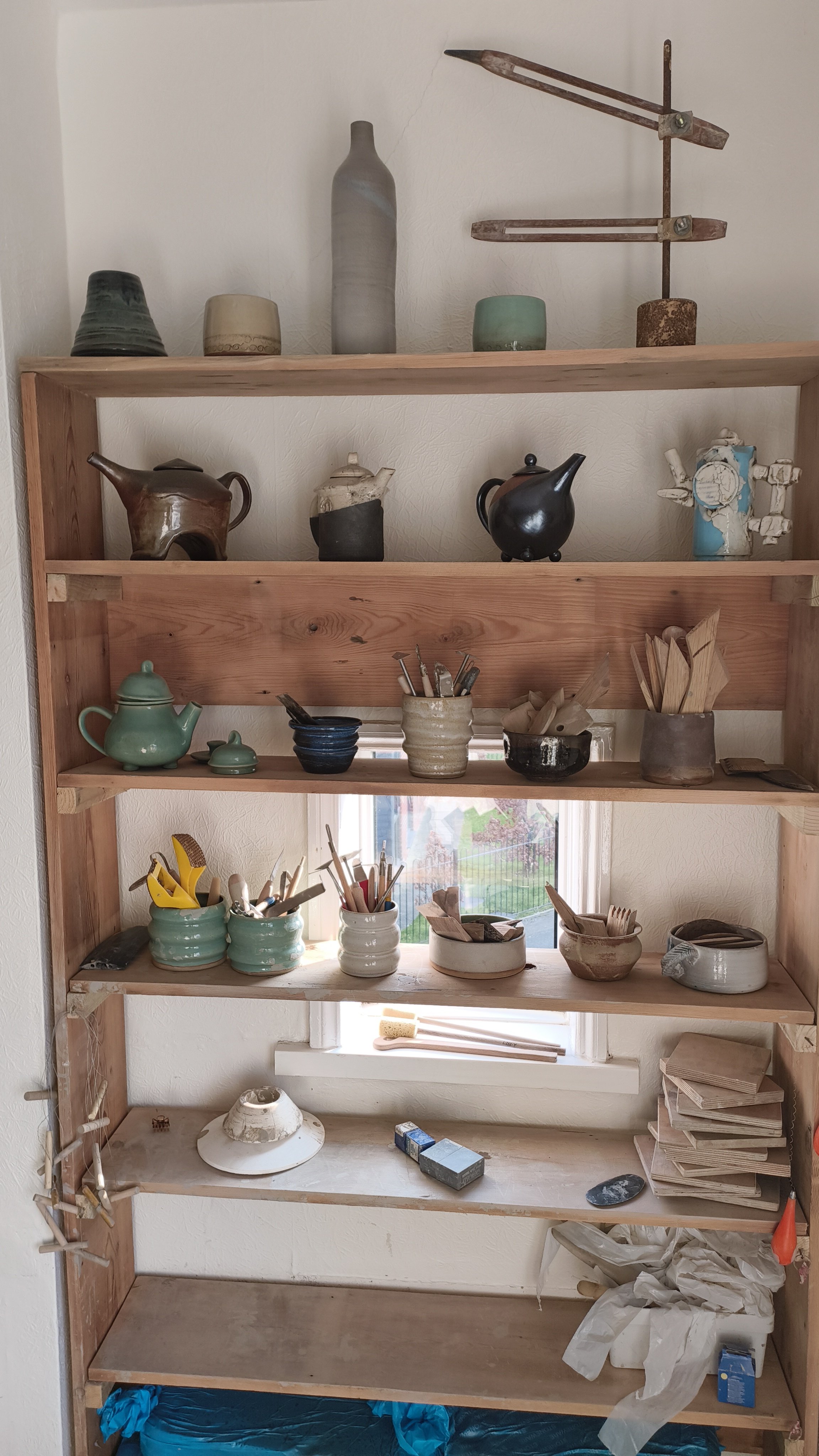 Wooden shelves displaying various pottery items, tools, and a stack of papers. Includes teapots, cups, and containers holding brushes and wooden utensils, with some ceramic materials and crafting supplies also visible.