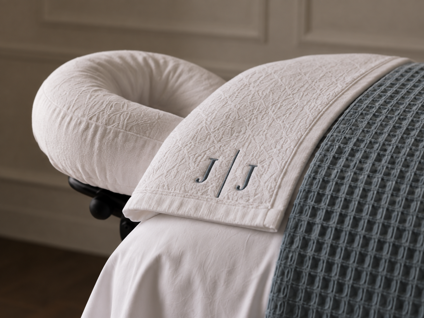 A white embroidered towel with the initials 'J J' draped over a bed, with a rolled white bolster pillow and a blue textured bedspread.