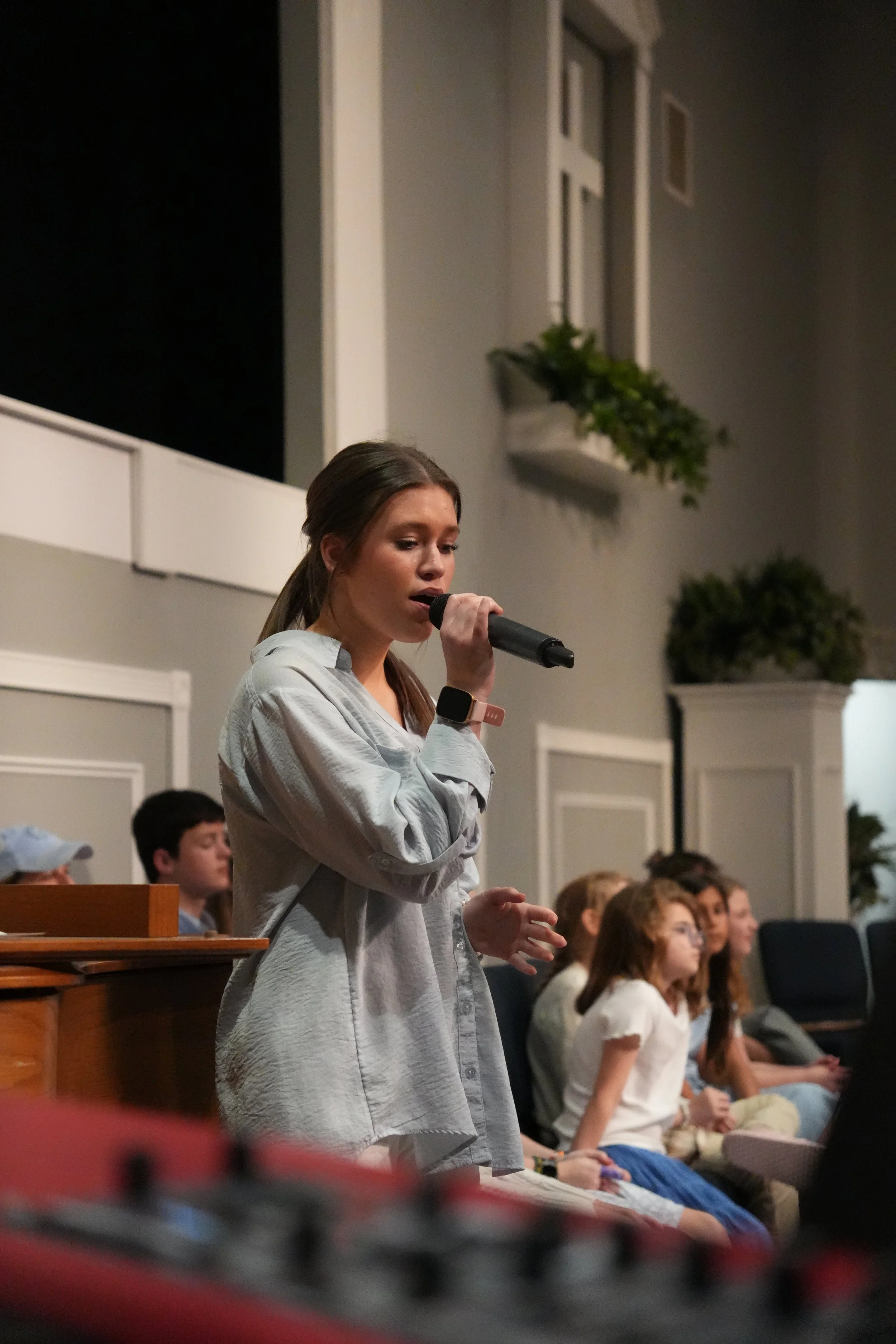 A young woman with long brown hair holding a microphone, speaking or singing in front of an audience in an indoor setting. Behind her, there are children seated, some wearing white dresses or shirts, and the room is decorated with plants and framed windows.