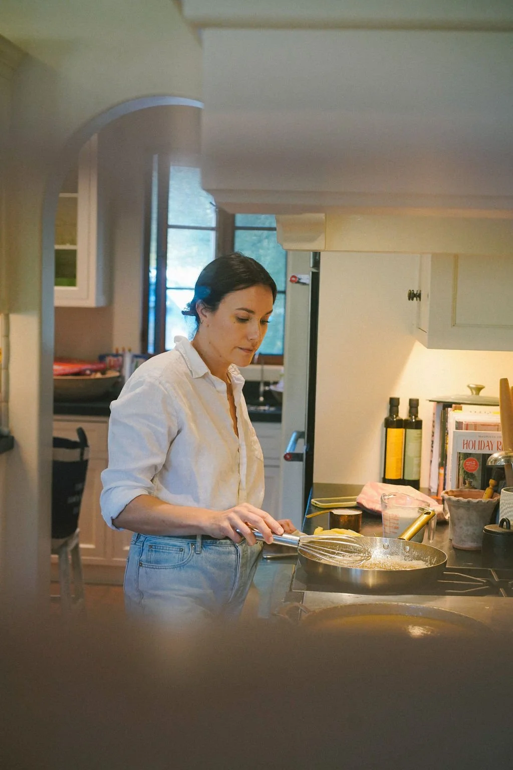 A woman cooking in a kitchen, standing at the stove with a frying pan, whisking ingredients.