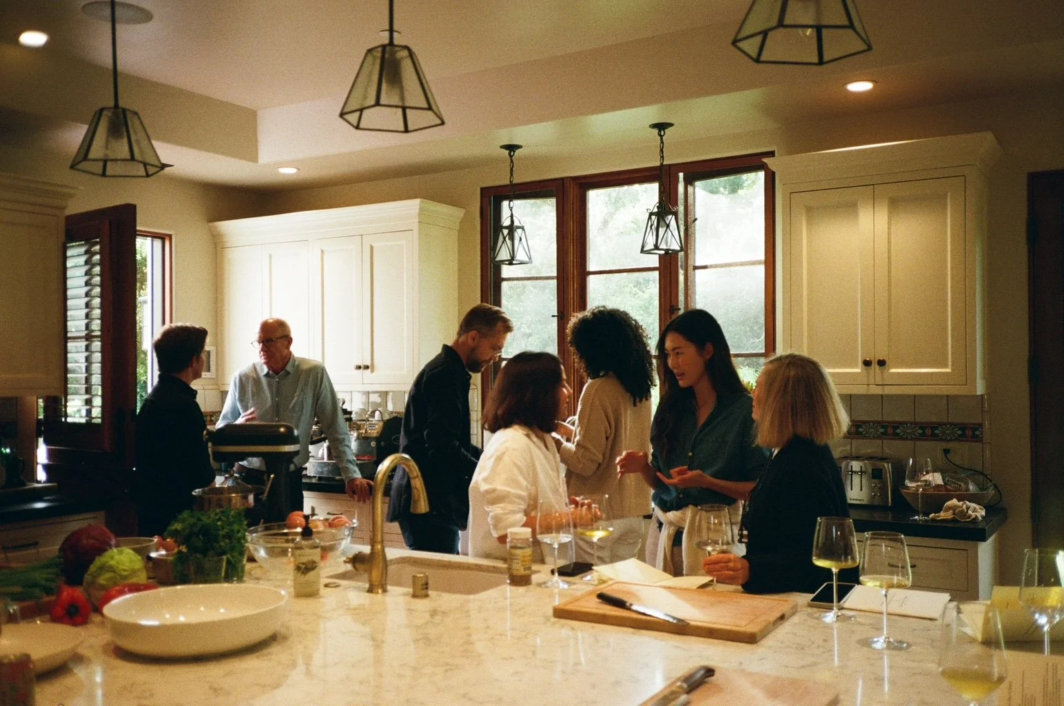 Group of people gathered in a kitchen, socializing and preparing food, with a modern design and ample natural light.