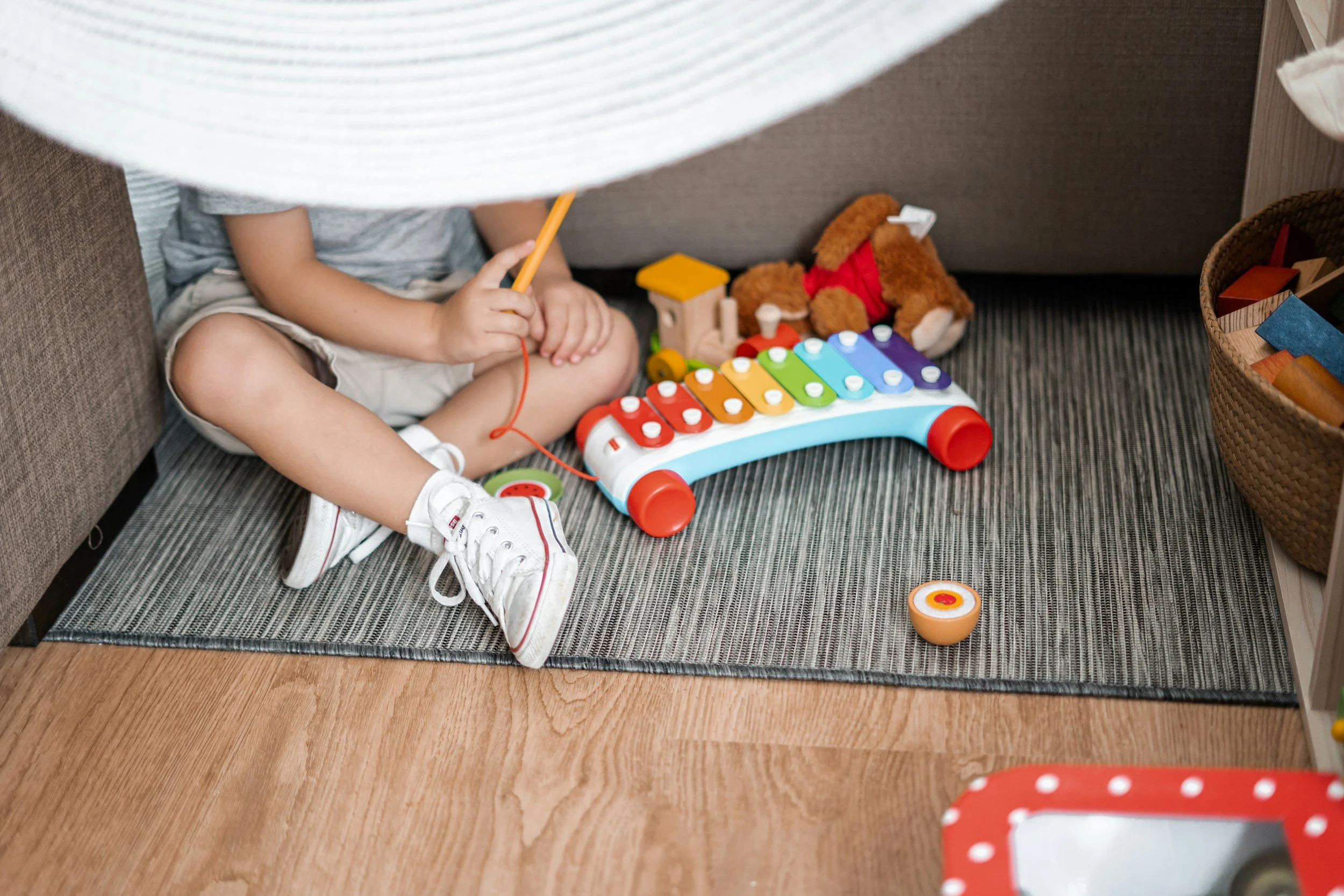Child sitting on a rug under a couch, playing with a colorful xylophone toy, surrounded by stuffed animals and storage baskets.