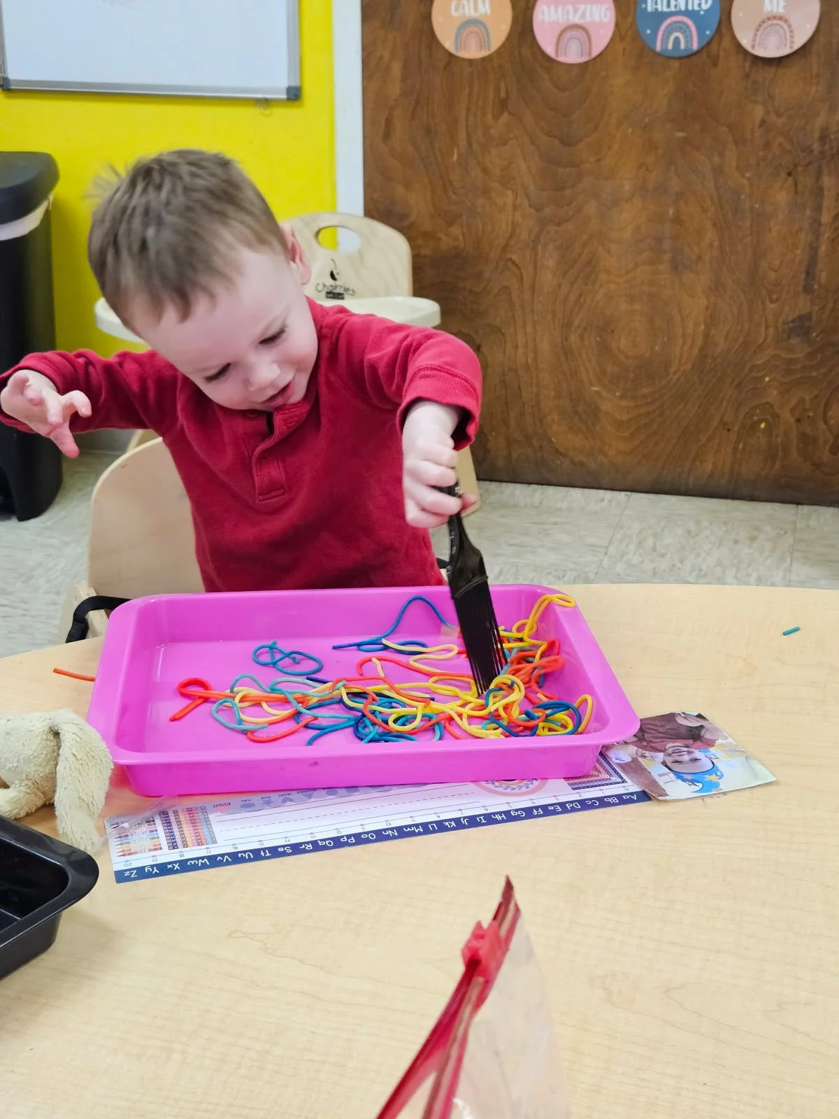 A young boy in a red long-sleeve shirt is playing with colorful rubber band bracelets in a pink tray at a classroom table.