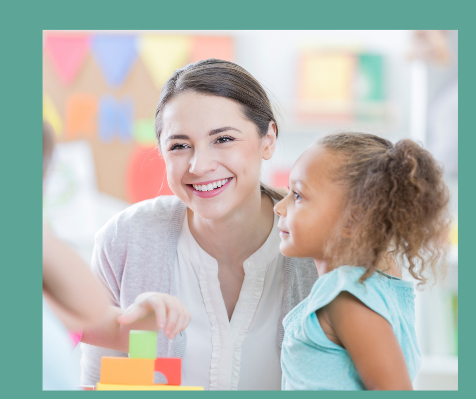 Teacher smiling with young child in classroom setting, with colorful blocks and decorations.