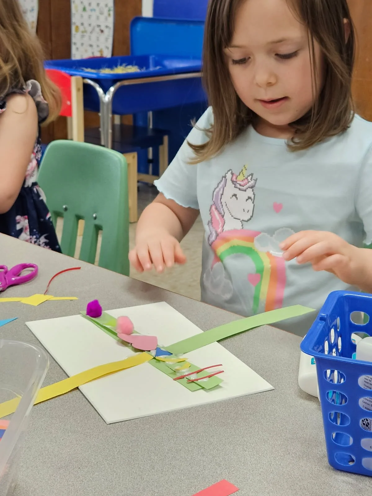 A young girl with shoulder-length brown hair wearing a unicorn T-shirt sitting at a table, working on a craft project with colorful paper and foam shapes. There are craft supplies and a blue storage bin on the table.