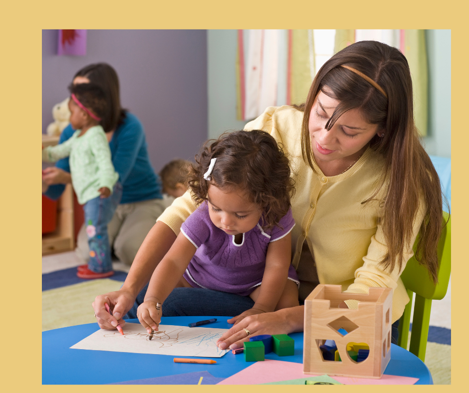 Teacher and child engaged in a drawing activity at a daycare, with other children and toys in the background.