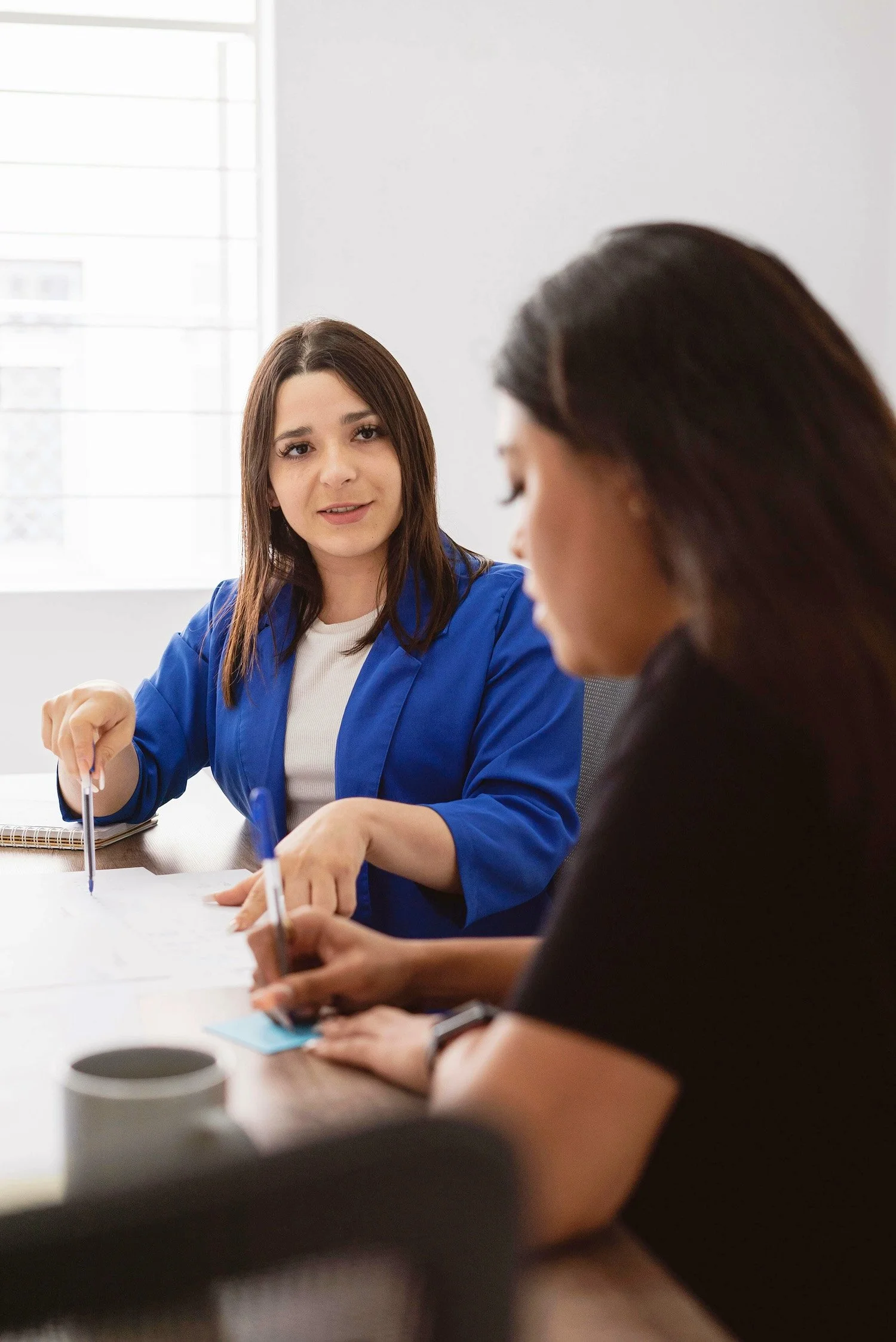 Two women are having a discussion at a conference table in a bright, modern office. One woman, with brown hair and a blue blazer, is speaking and gesturing with a pen while the other woman, with dark hair, listens attentively.