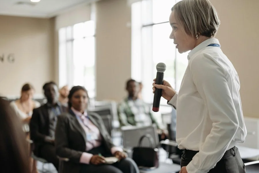 A woman in business attire giving a presentation to a group of diverse seated attendees in a conference room.