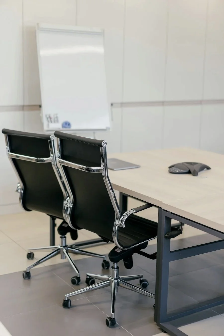 An empty office conference room with two black and chrome office chairs, a white table, a conference phone, and a whiteboard on the wall.