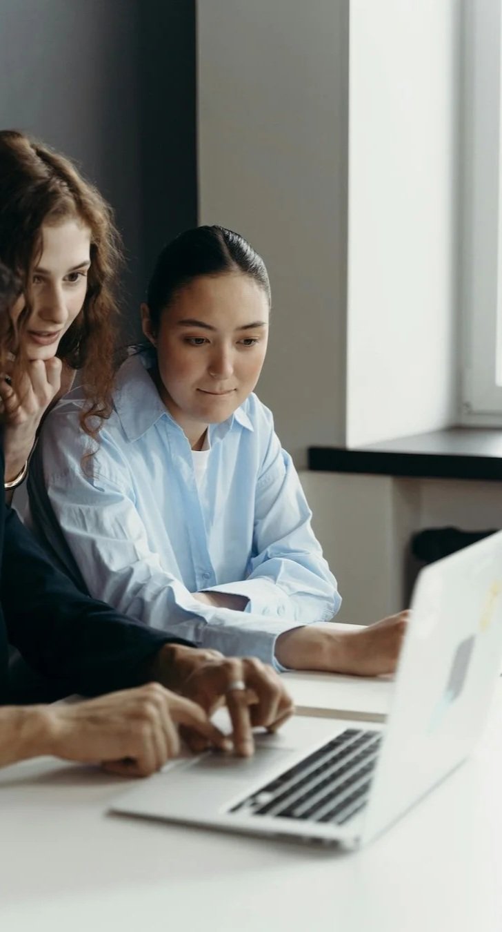 Two women looking at a laptop screen, one with curly hair and the other with dark straight hair, in an office setting.