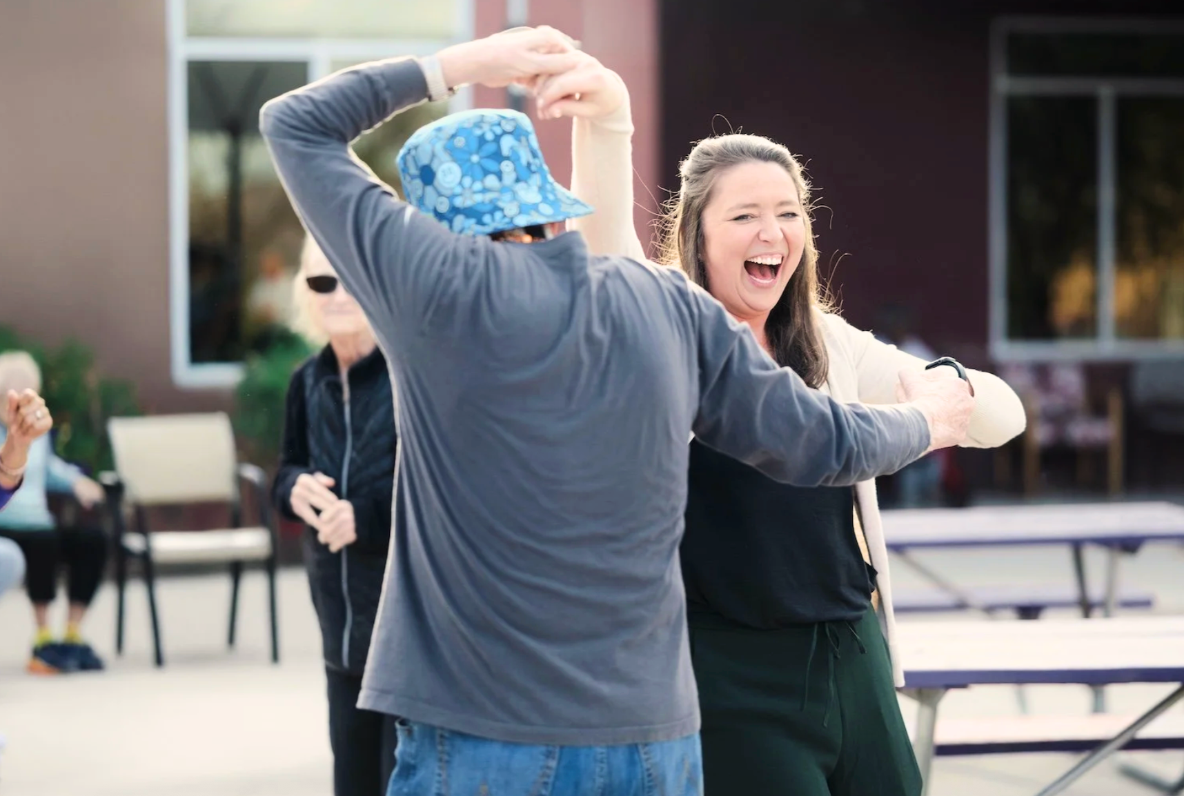 A woman with long brown hair and a black watch smiling and laughing as she dances with a man wearing a blue floral hat. Other people are seated and standing in the background outdoors.