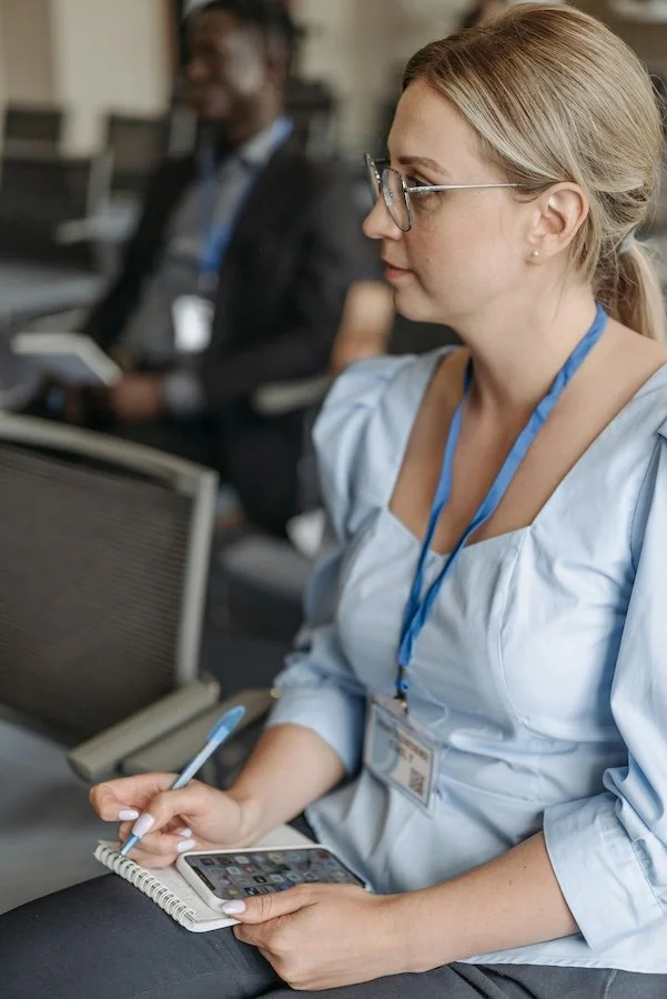 A woman with blonde hair, glasses, and a light blue shirt is sitting, holding a smartphone in one hand and a pen and notepad in the other. She is wearing a lanyard with an ID badge. In the background, there is a man in a suit blurred out.