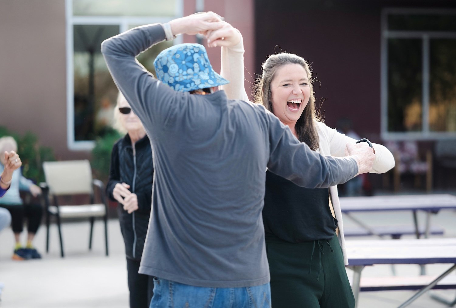 Two people are dancing and laughing outdoors near tables. One person wears a blue hat with a floral pattern, gray hoodie, and jeans. The other is a woman with long brown hair, wearing a light-colored sweater and black pants.