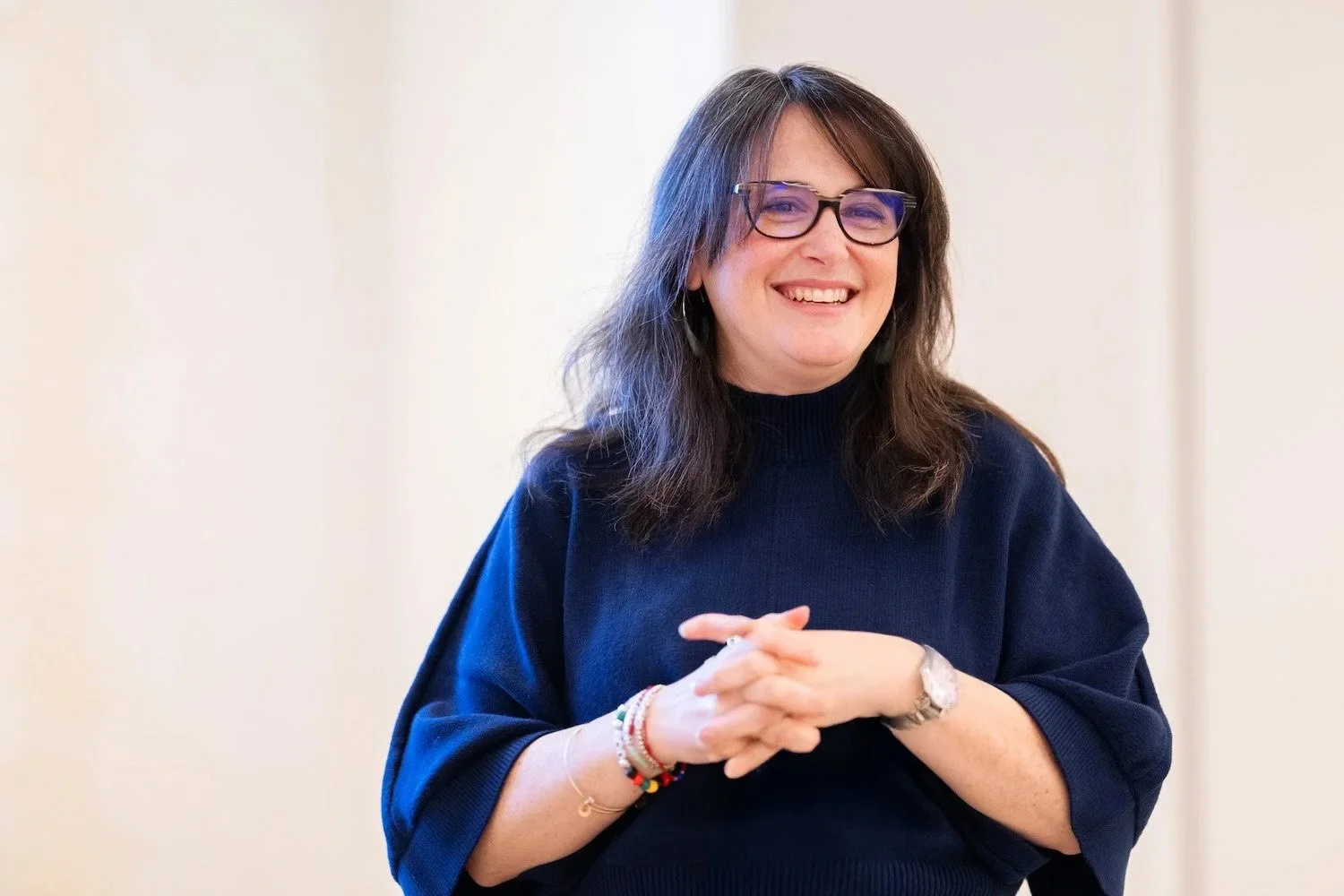 A woman with dark brown hair, wearing glasses, a navy blue sweater, and various bracelets, smiling and standing against a plain background.