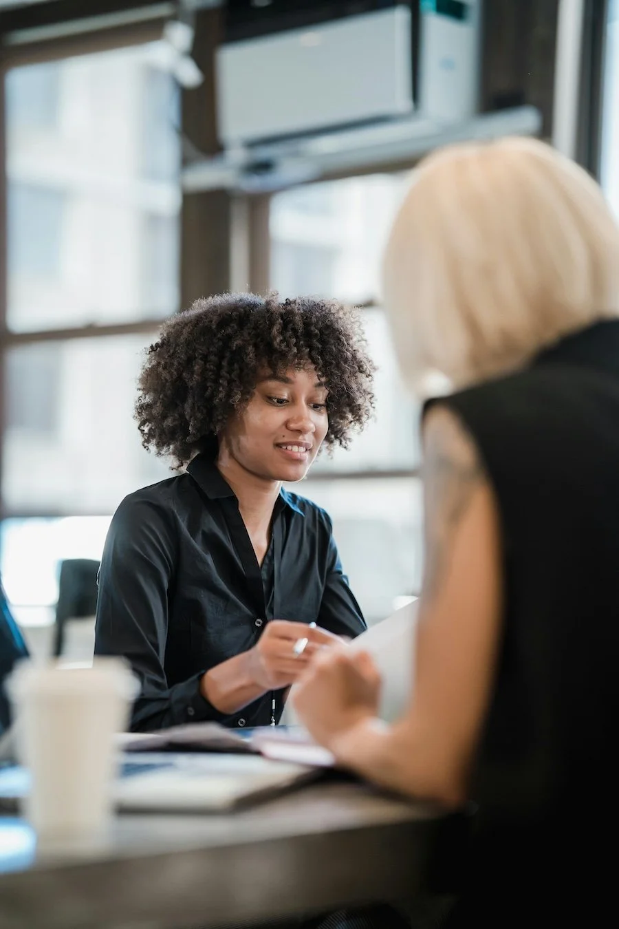 Two women in an office, one with curly hair holding a document and the other with blonde hair and tattoos on her arm, engaged in a discussion at a table.