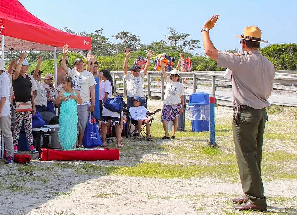 A park ranger leading an outdoor activity with a group of people under a red canopy tent. Participants are raising their hands, suggesting interaction or agreement. The setting appears to be a park with green grass and trees.