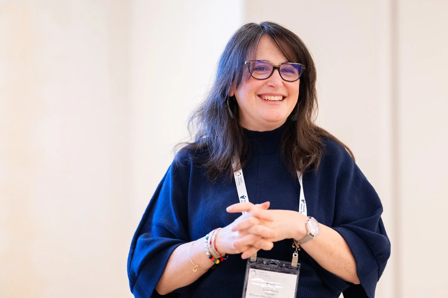 A woman with shoulder-length brown hair, glasses, and a navy blue sweater, smiling with her hands clasped, wearing a name badge and colorful bracelets, standing in a well-lit indoor setting.