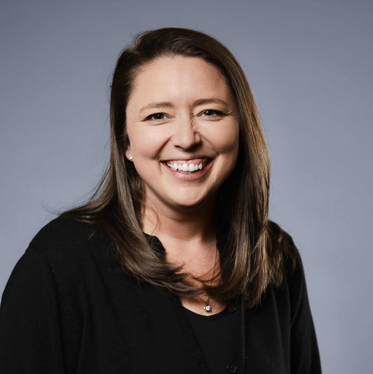 A woman smiling against a plain blue background, wearing a black top and a necklace with a pendant.