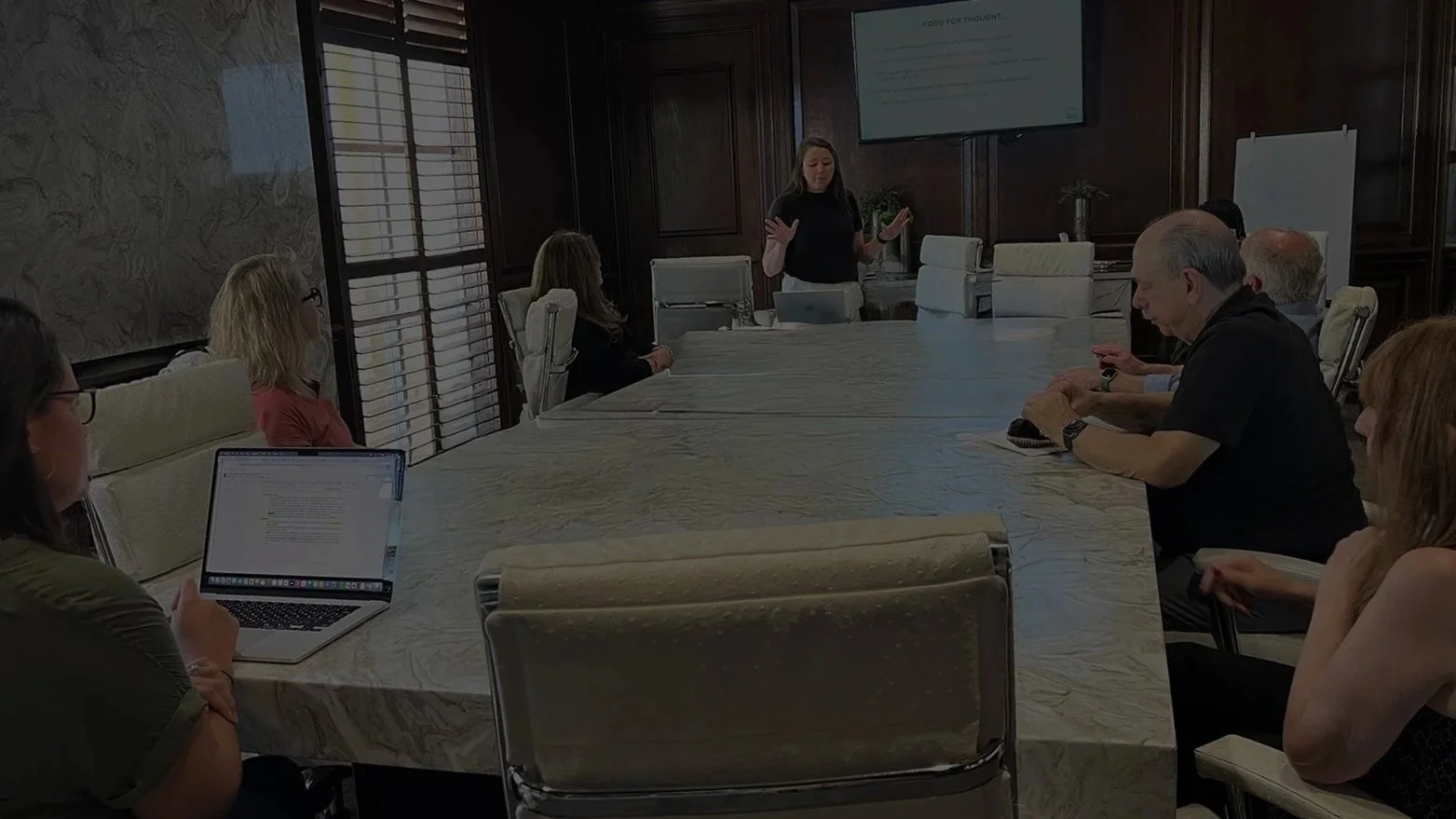 A woman giving a presentation in a conference room with several seated attendees, some using laptops and one using a phone.
