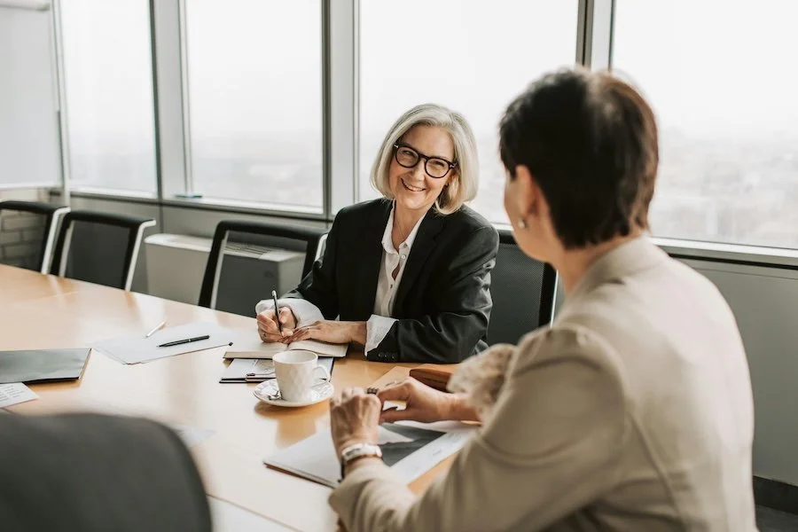 Two professional women having a meeting in a conference room with large windows, paperwork and coffee on the table, one woman smiling and taking notes.