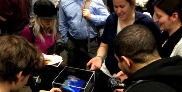 Group of people gathered around a small holographic display, some holding controllers, in an indoor setting.