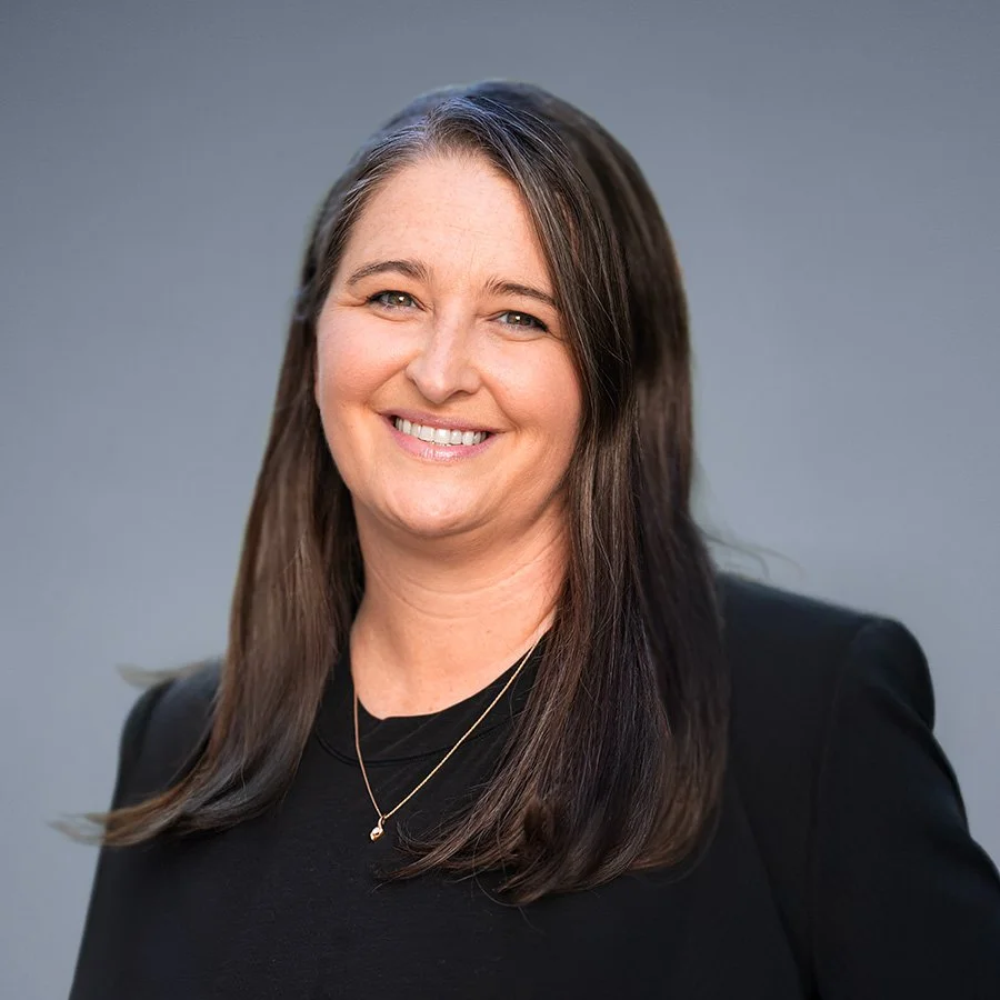 A smiling woman with long brown hair wearing a black top and a gold necklace.