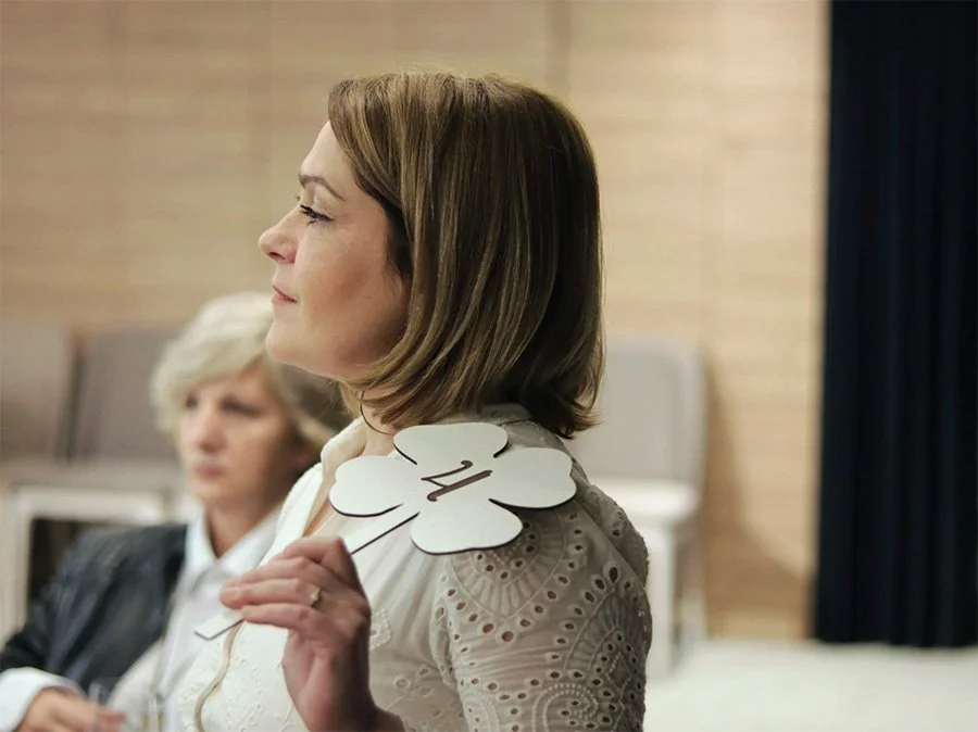 A woman with shoulder-length hair holding a paper cutout of a four-leaf clover with the number 24 on it, in a room with a blurred background featuring another woman and some furniture.